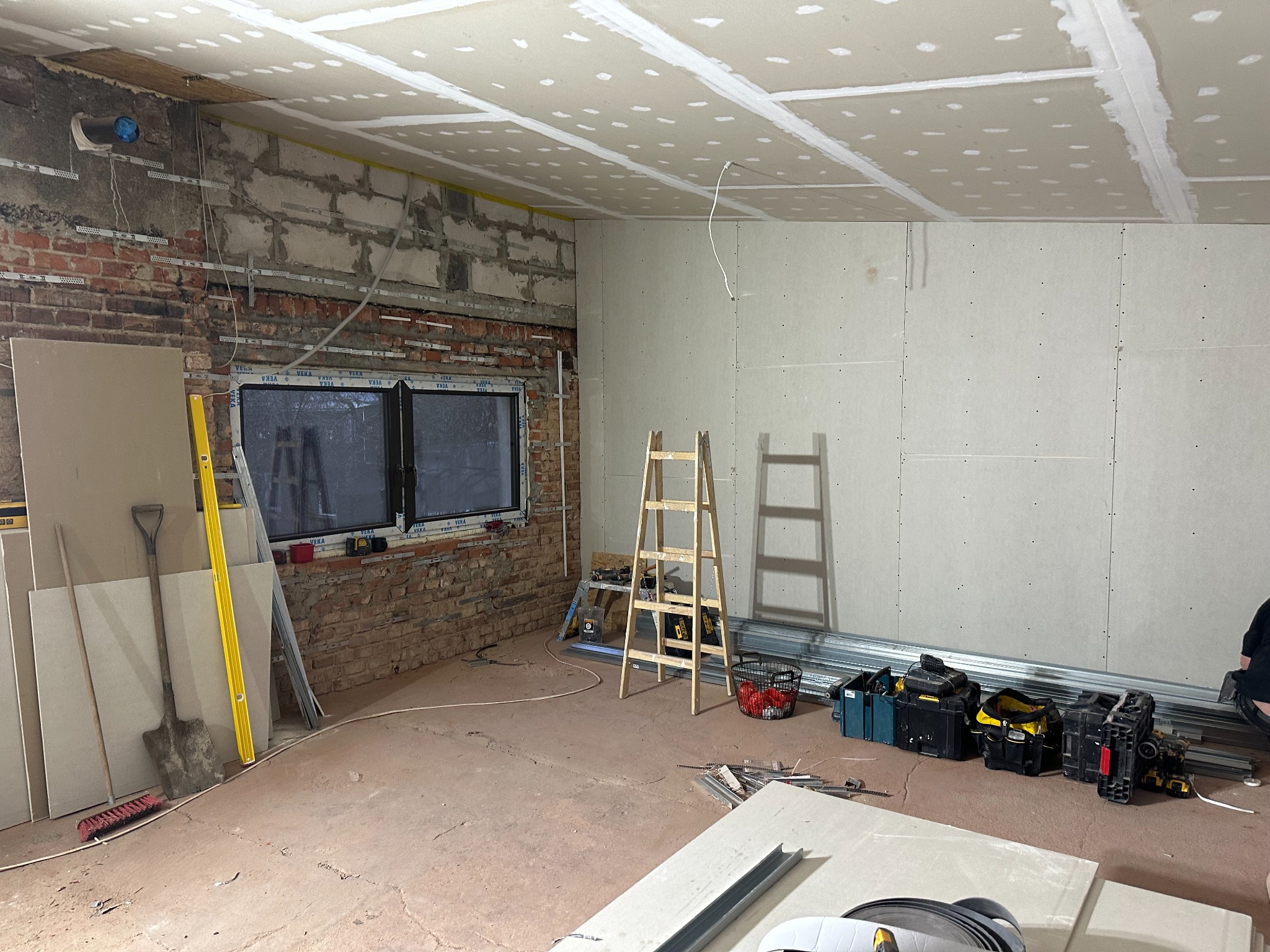 Wnętrze w trakcie prac w Łodzi: widoczna surowa cegła, okno z PCV, częściowo zamontowana płyta na suficie i ścianach, drabiny, narzędzia i materiały budowlane.