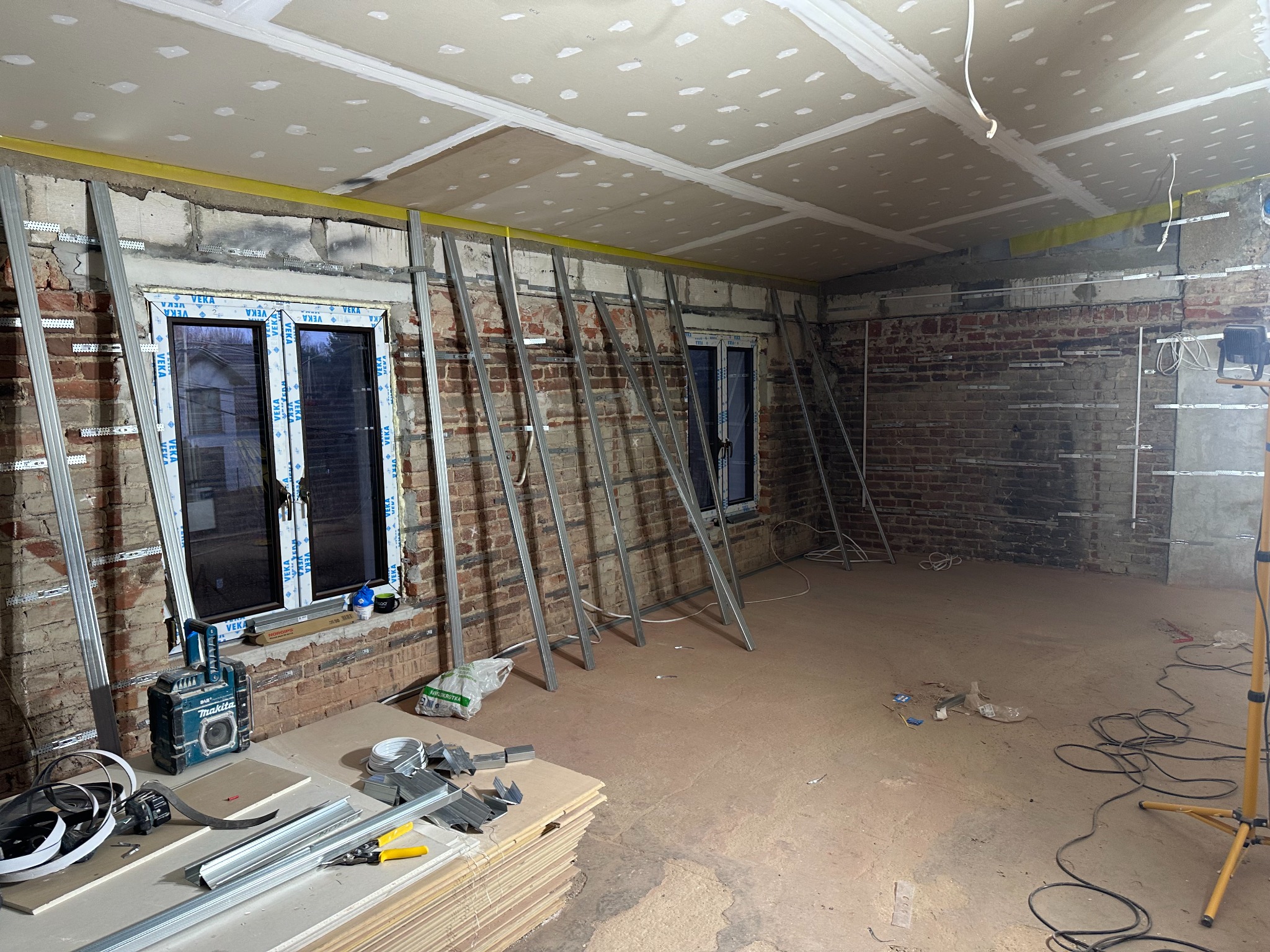 Wnętrze w trakcie prac remontowych: widoczne metalowe profile konstrukcyjne przy ceglanej ścianie, częściowo zamontowane płyty na suficie, narzędzia budowlane i radio Makita na podłodze, okna...