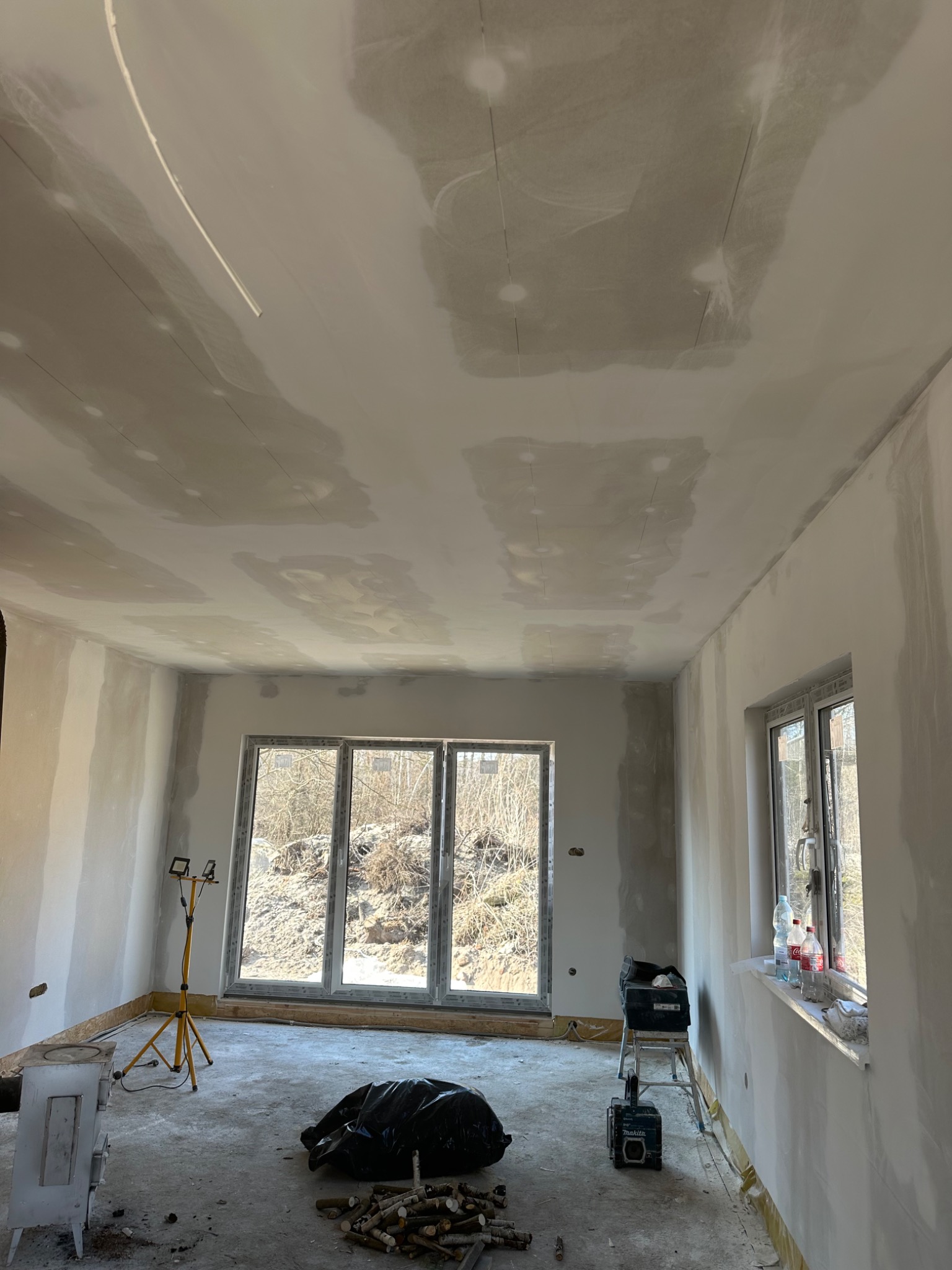 Wnętrze w trakcie prac wykończeniowych w Łodzi: surowe ściany i sufit po wstępnym wyrównaniu, widoczne ślady po szpachlowaniu, narzędzia pracy i prowizoryczne ogrzewanie kominkowe.