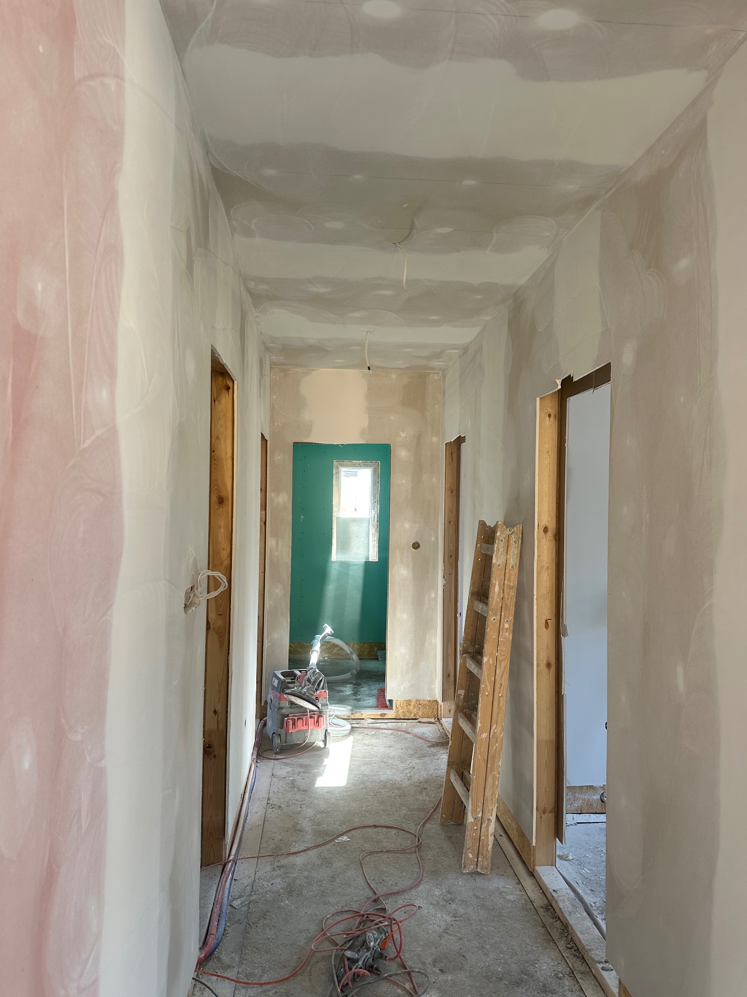 Korytarz w trakcie prac wykończeniowych: ściany częściowo pokryte gładzią, widoczna drabina, szlifierka do gładzi i przewody na podłodze, w tle pomalowane na zielono pomieszczenie z oknem.