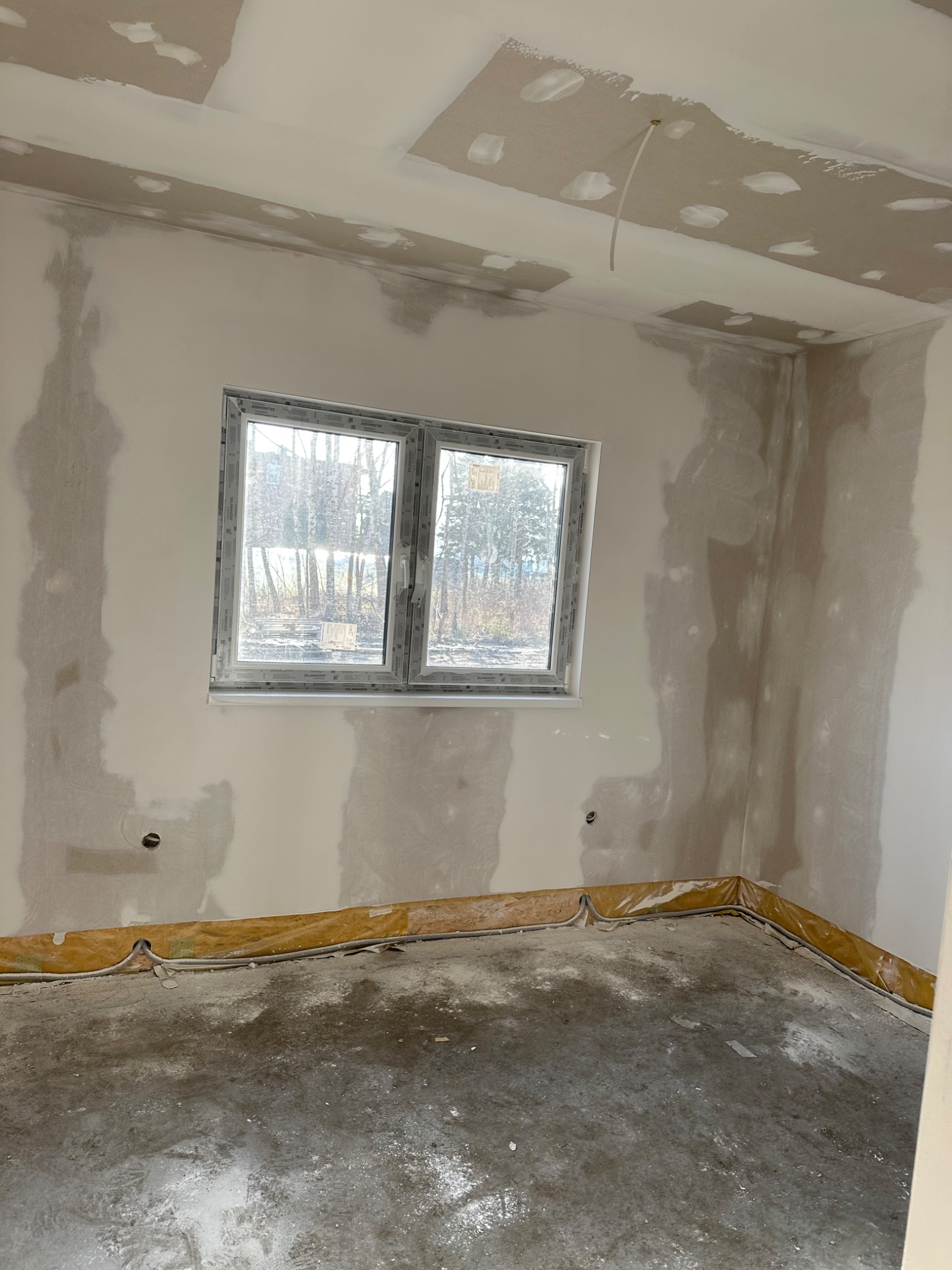 Pokój w trakcie prac wykończeniowych: ściany z widocznymi punktowymi poprawkami i przygotowaniem pod malowanie, surowa podłoga, okno z widokiem na drzewa, widoczne zabezpieczenia podłogi przy...