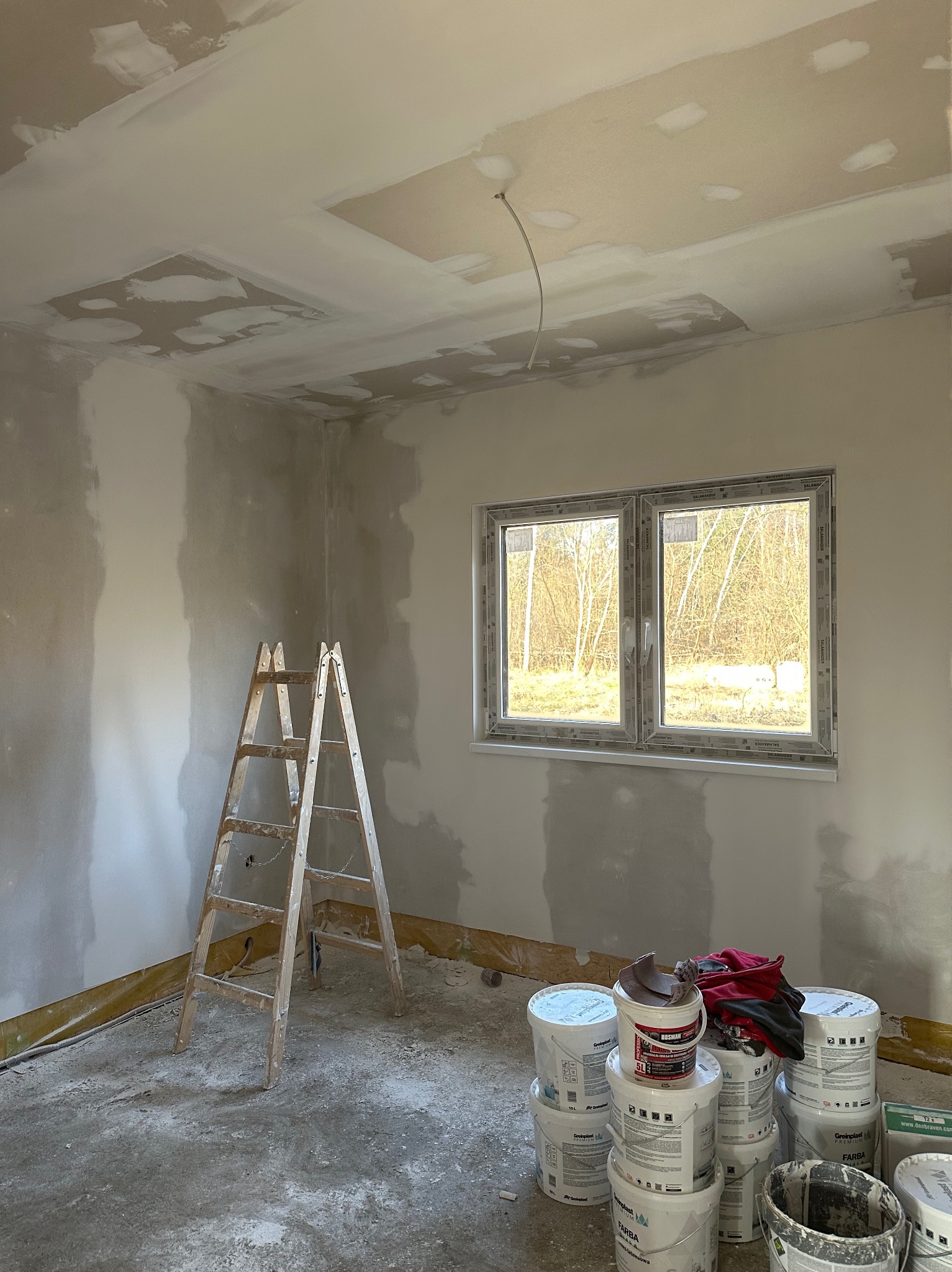 Pokój w trakcie prac wykończeniowych: ściany przygotowane pod malowanie, częściowo wyszpachlowane, widoczne wiaderka z farbą, drabina drewniana i okno z widokiem na drzewa.