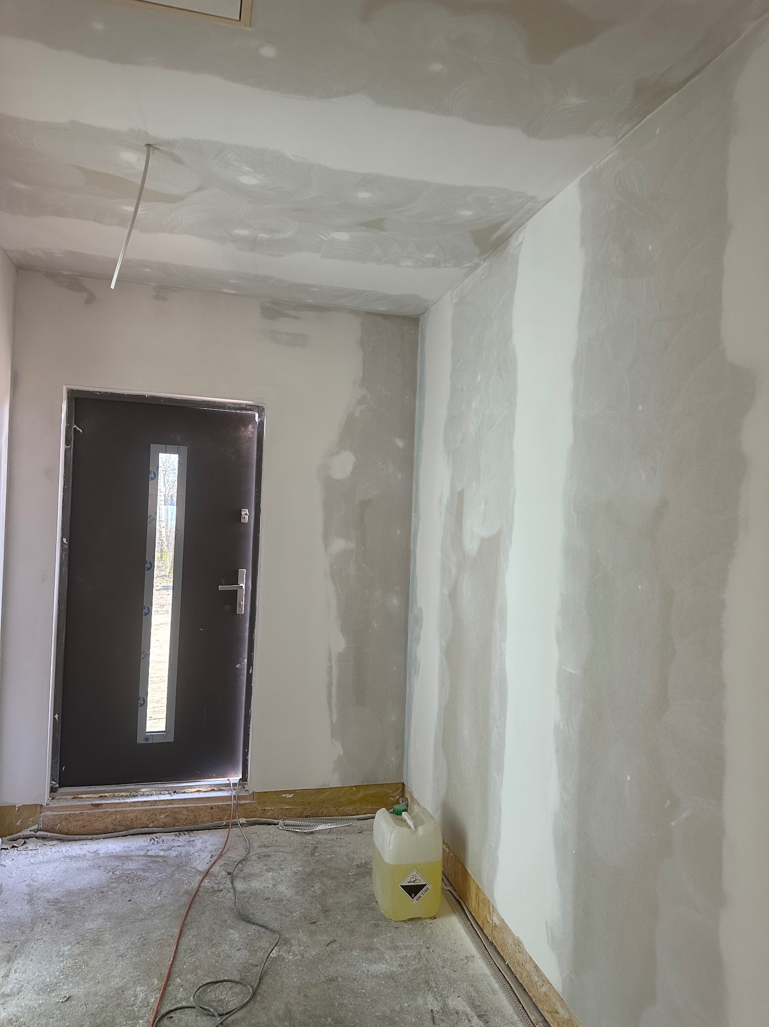 Wnętrze w trakcie prac wykończeniowych: ściany częściowo pokryte gładzią, widoczna surowa podłoga, drzwi wejściowe i kanister z płynem na podłodze.