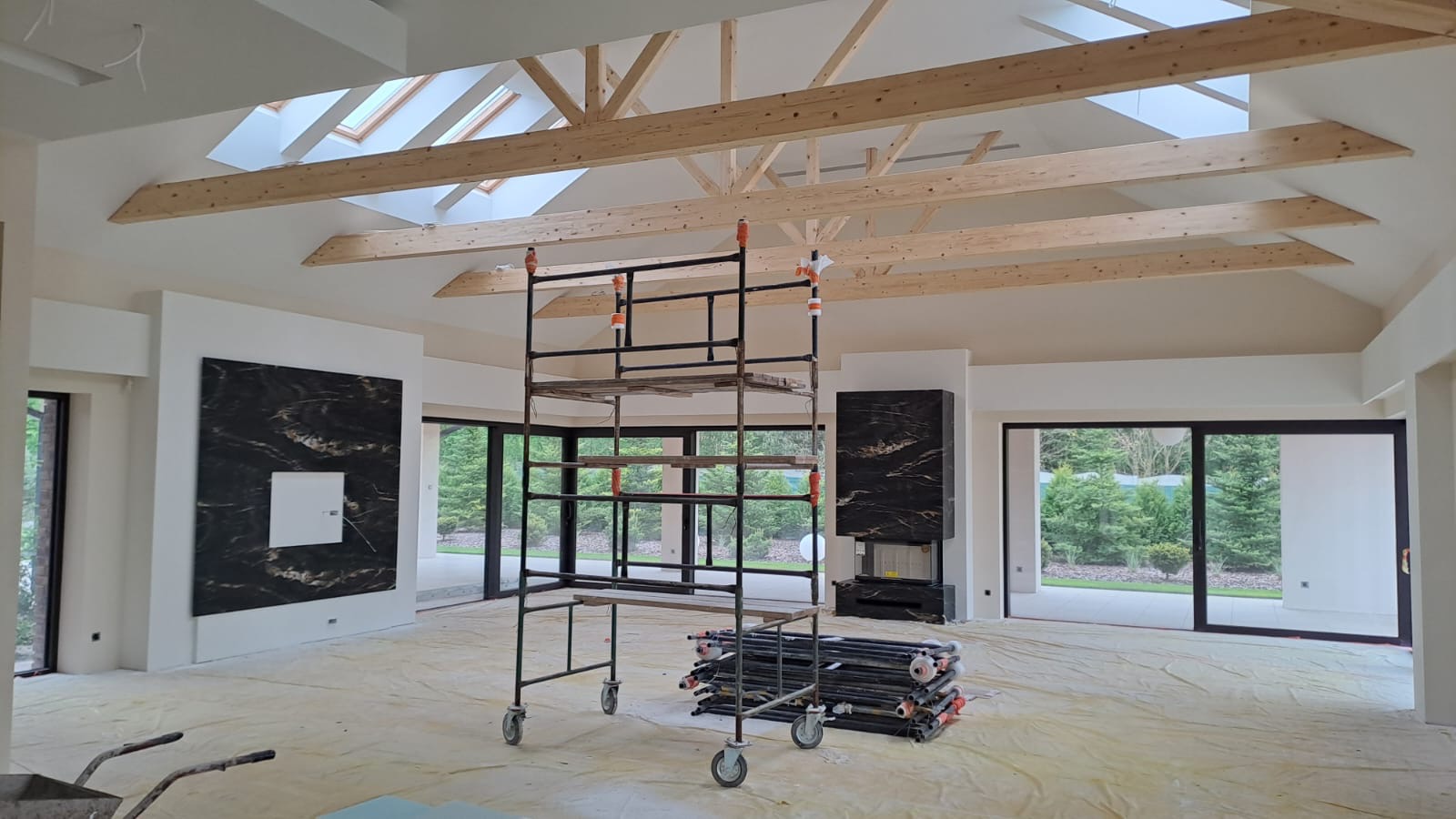 Wnętrze w trakcie prac wykończeniowych: widoczne drewniane belki stropowe, rusztowanie na kółkach, zabezpieczona folią podłoga oraz czarne panele dekoracyjne na ścianach.