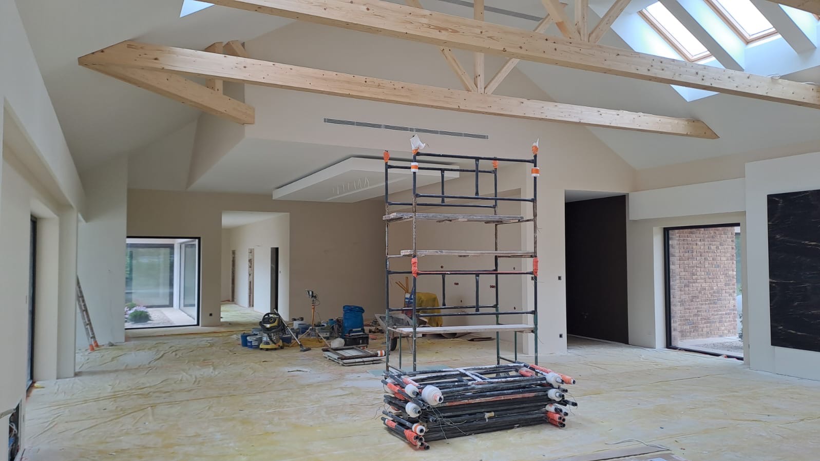 Wnętrze w trakcie prac wykończeniowych: widoczne drewniane belki stropowe, rusztowanie, zabezpieczona podłoga oraz narzędzia malarskie i szlifierka.