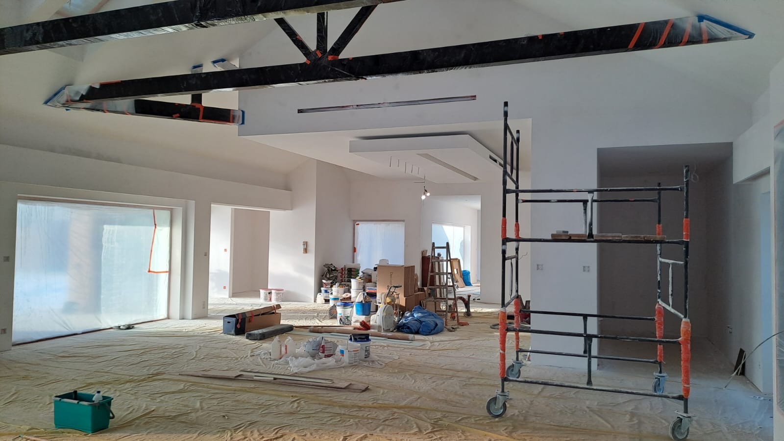 Wnętrze domu w trakcie prac wykończeniowych, z zabezpieczoną podłogą, rusztowaniem, przygotowanymi farbami i widocznymi elementami konstrukcyjnymi sufitu.
