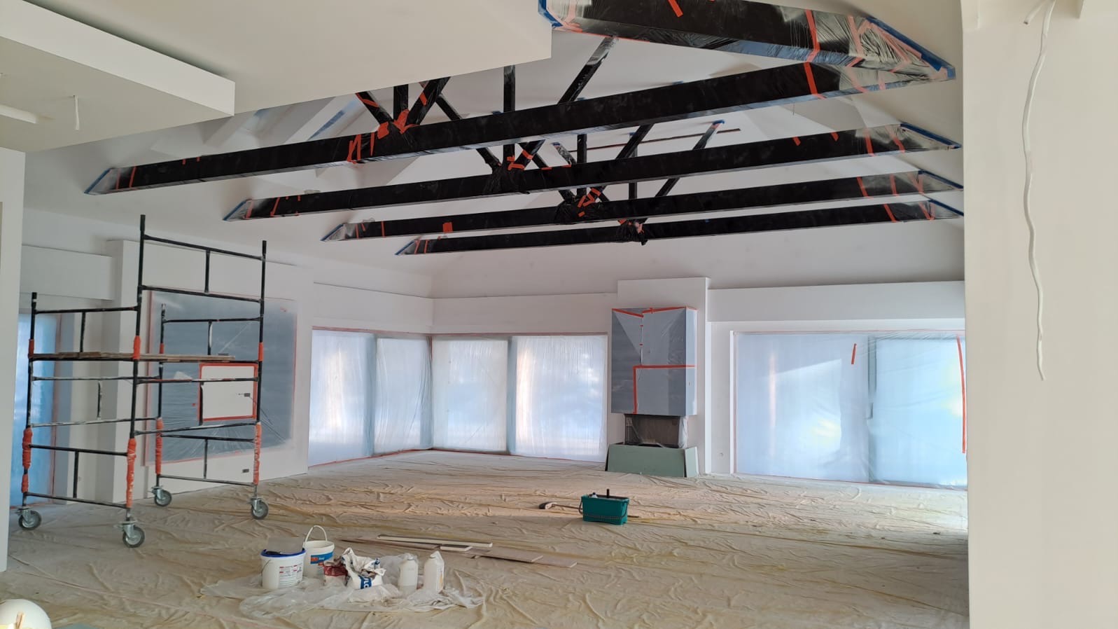 Wnętrze w trakcie prac przygotowawczych: podłoga i okna zabezpieczone folią malarską, widoczne rusztowanie, czarne elementy konstrukcyjne pod sufitem z zabezpieczeniami, pojemniki z farbą.