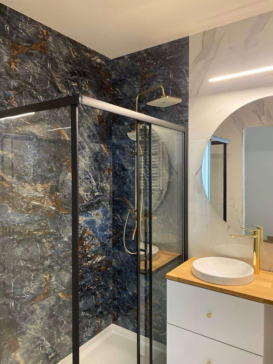 Nowoczesna łazienka z kabiną prysznicową z czarnymi ramami, ściany wyłożone płytkami imitującymi marmur w odcieniach granatu i złota, oraz białą umywalką nablatową z baterią w kolorze złota.