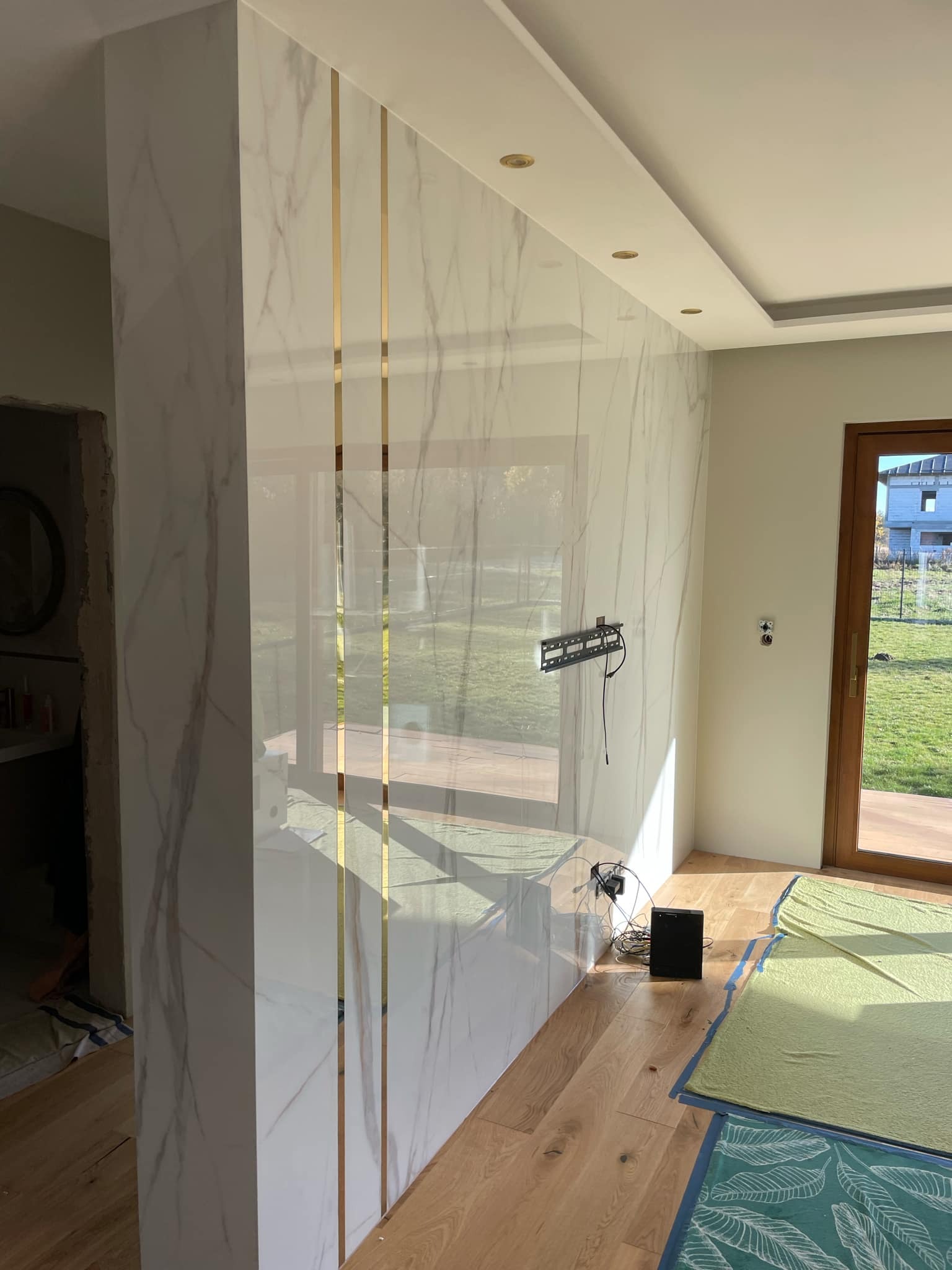 Ściana wykończona białym marmurem z złotymi listwami dekoracyjnymi, widoczny uchwyt na telewizor i fragment podłogi zabezpieczony folią malarską.