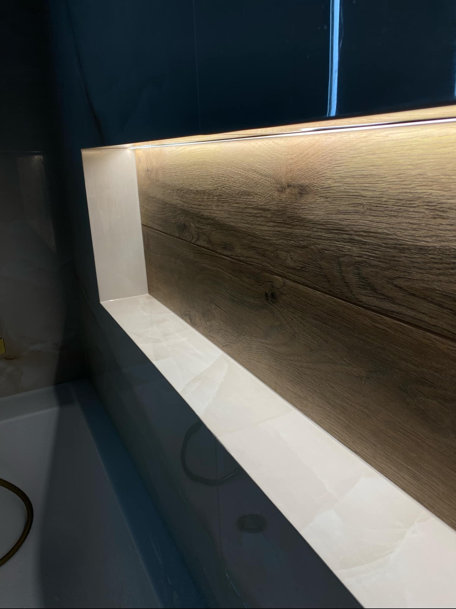 Wnęka w łazience wykończona płytkami imitującymi drewno i marmur, podświetlona listwą LED, widoczny fragment wanny.