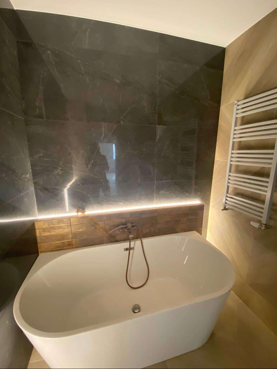 Biała wanna wolnostojąca z brązową armaturą na tle ściany z ciemnego marmuru z podświetleniem LED i grzejnikiem drabinkowym.