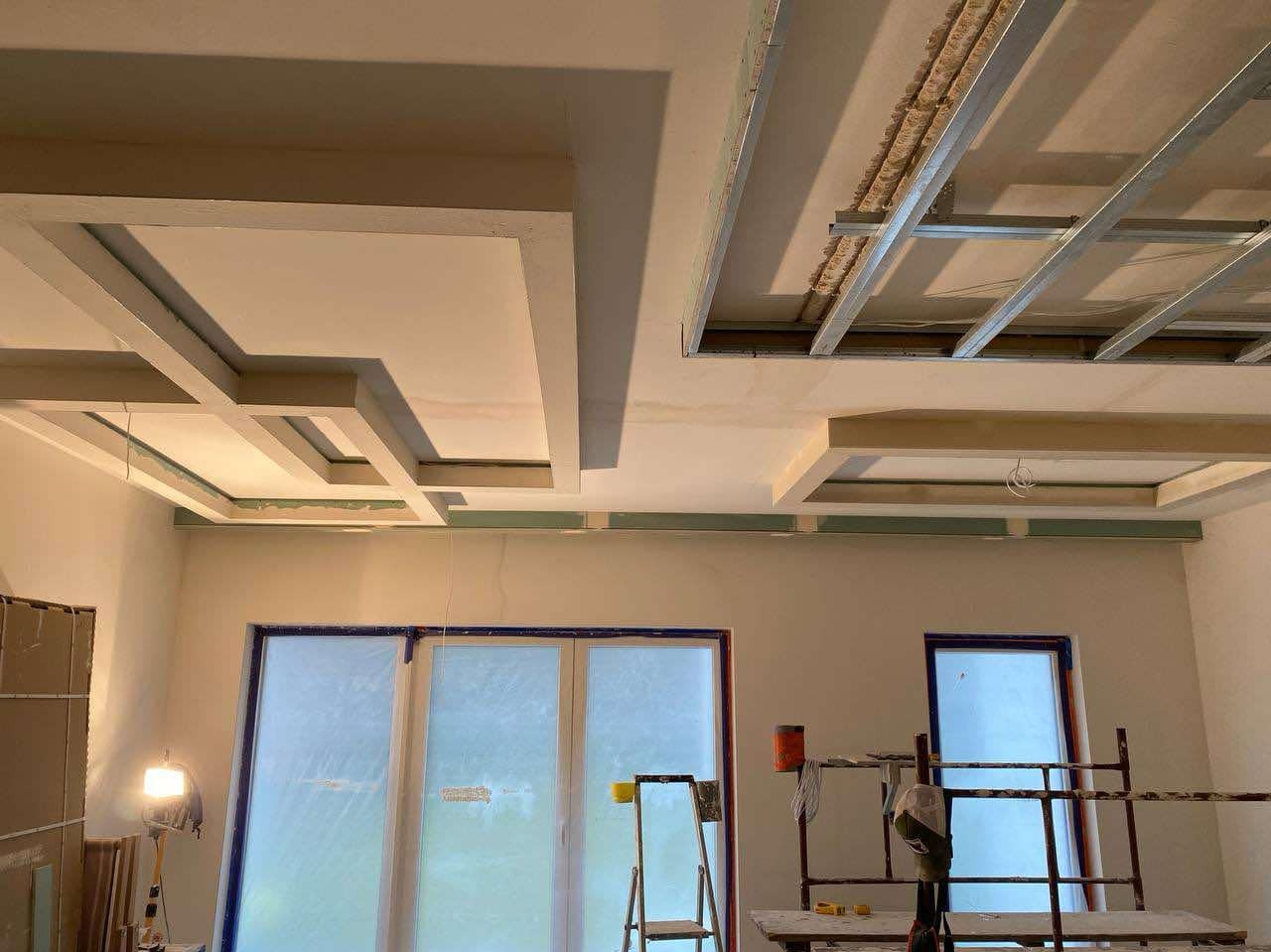 Wnętrze w trakcie prac wykończeniowych, widoczna konstrukcja sufitu podwieszanego z płyt gipsowo-kartonowych oraz rusztowanie i zabezpieczone okna folią.