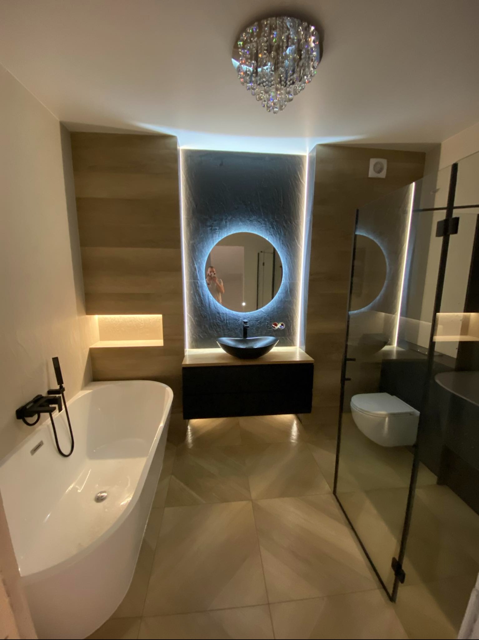 Nowoczesna łazienka z wolnostojącą białą wanną, czarną armaturą, okrągłym podświetlanym lustrem nad czarną szafką z umywalką typu vessel oraz kabiną prysznicową ze szklaną ścianą.