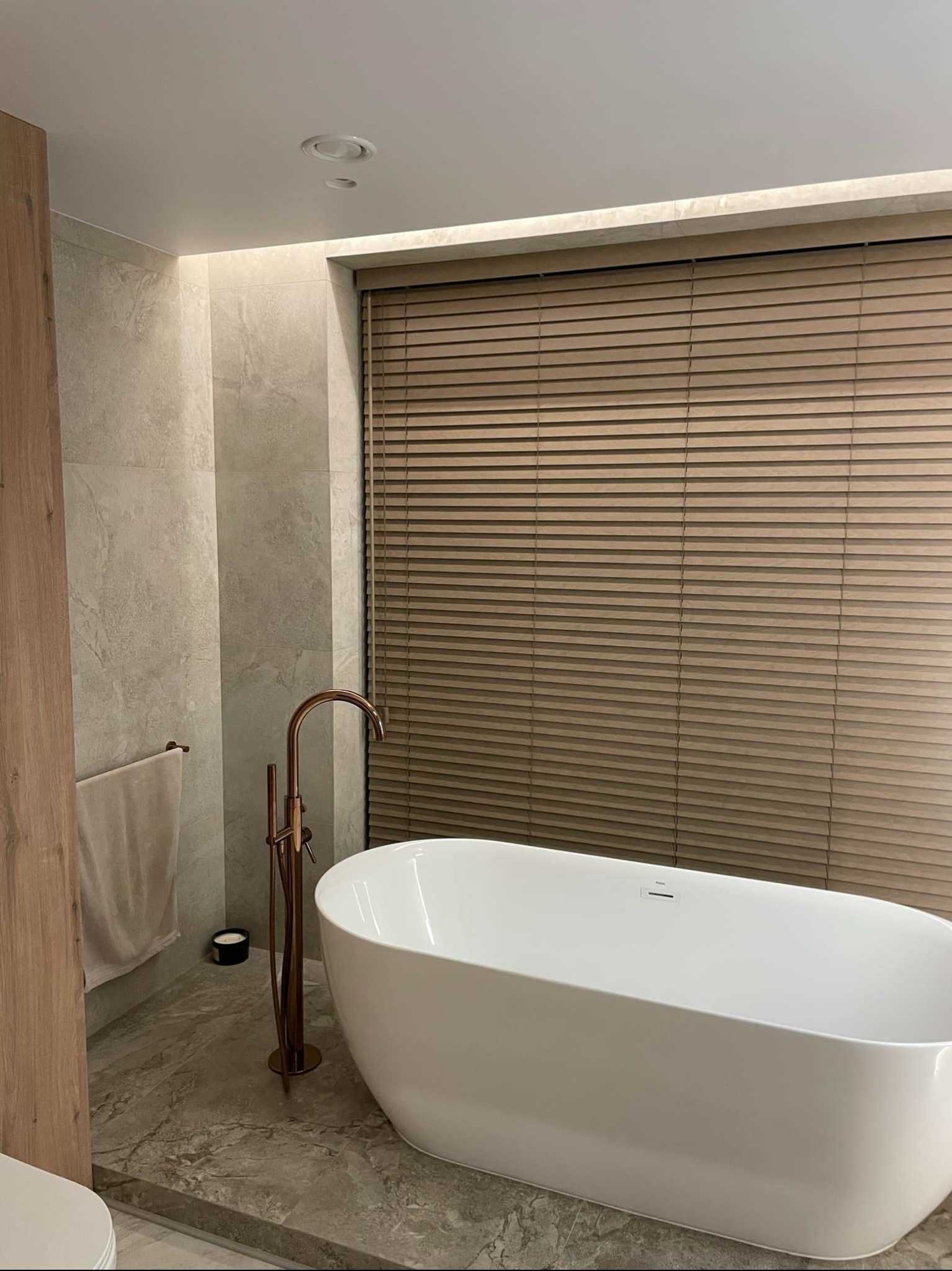 Nowoczesna łazienka z wolnostojącą białą wanną, złotą armaturą i drewnianymi żaluzjami w oknie.