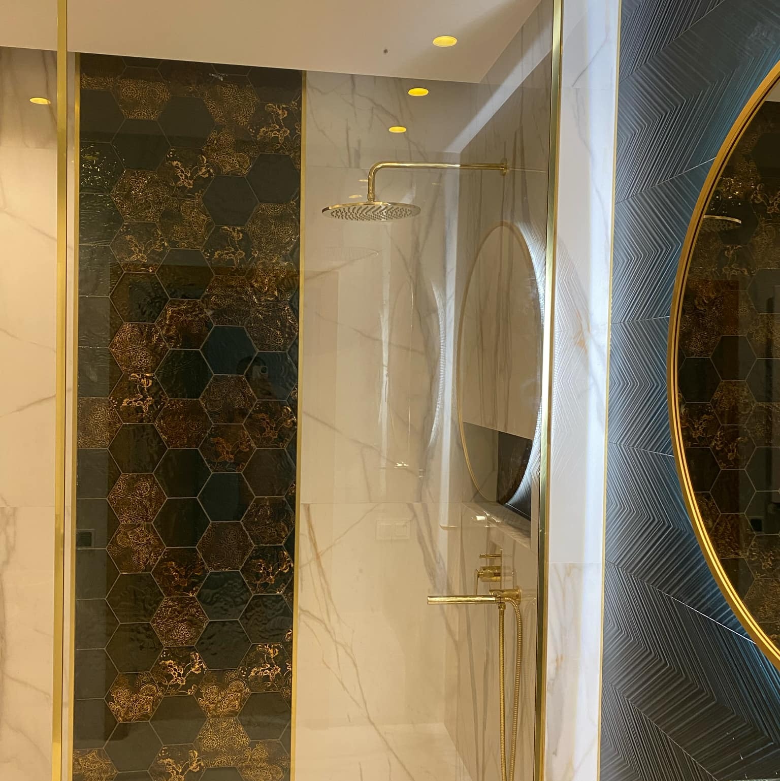Nowoczesna kabina prysznicowa z deszczownicą, wykończona płytkami heksagonalnymi w odcieniach czerni i złota, marmurowymi panelami oraz panelami z geometrycznym wzorem, całość podkreślona złotymi...
