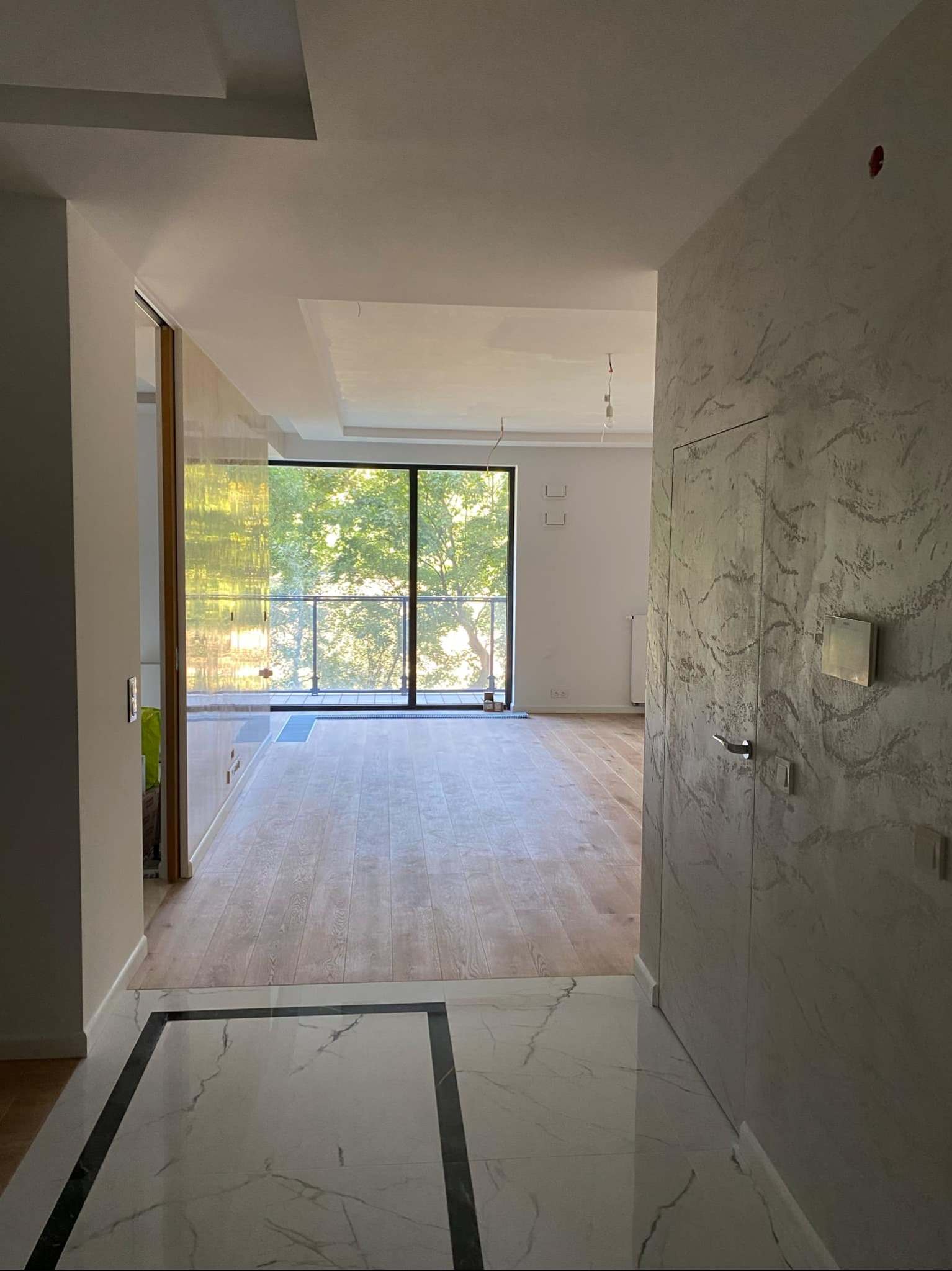 Wnętrze mieszkania w trakcie wykańczania: parkiet, marmurowe płytki podłogowe z czarnymi obramowaniami, ściana z dekoracyjnym tynkiem w odcieniach szarości i ukryte drzwi.