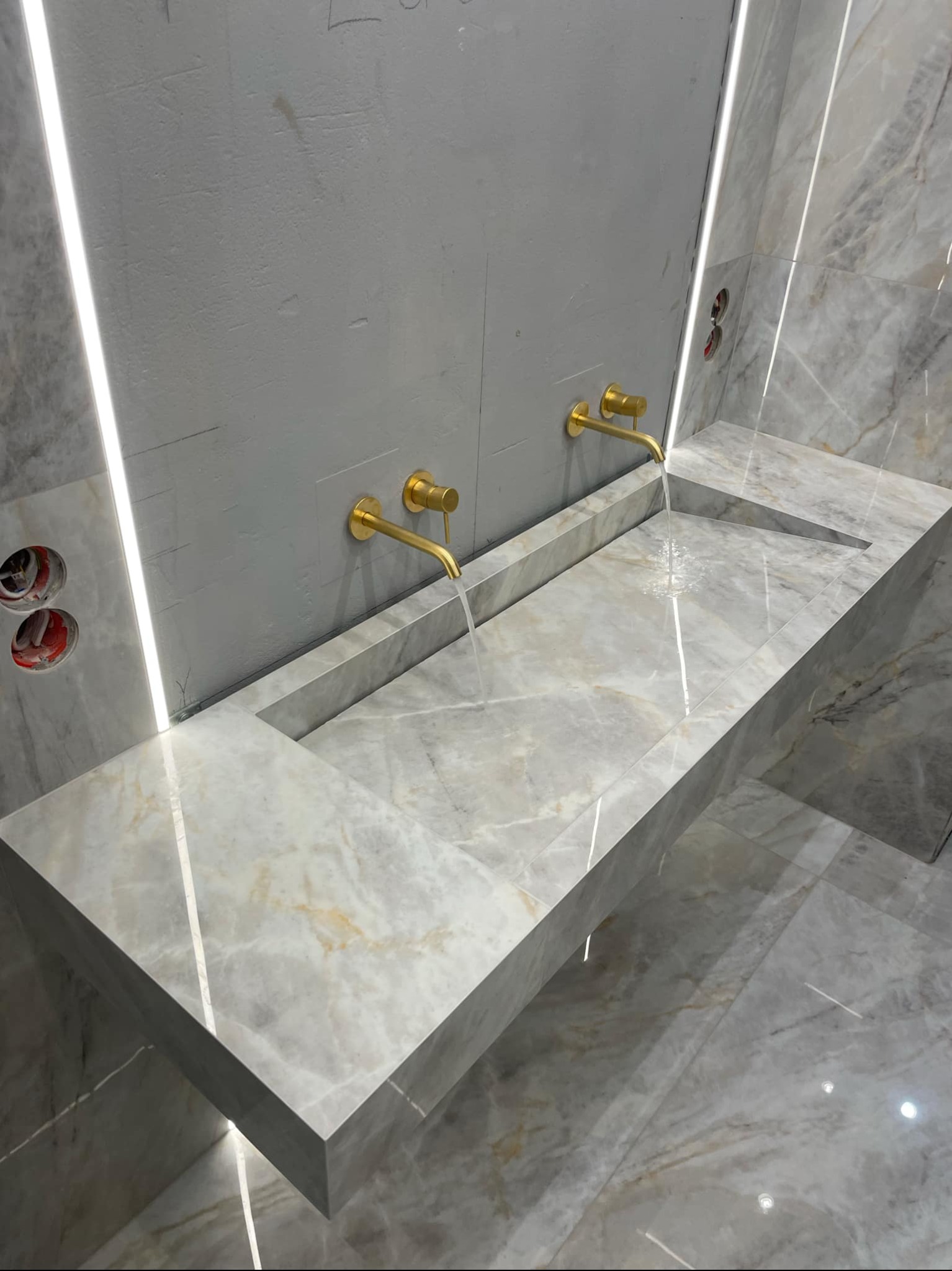 Nowoczesna łazienka z marmurową umywalką, złotą armaturą i podświetleniem LED, widoczne otwory pod gniazdka elektryczne.