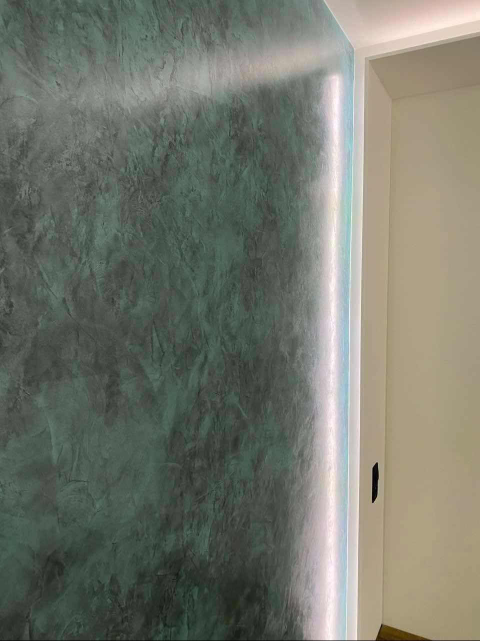 Ściana z dekoracyjnym tynkiem w odcieniach zieleni i szarości, oświetlona pionową listwą LED przy framudze drzwi.