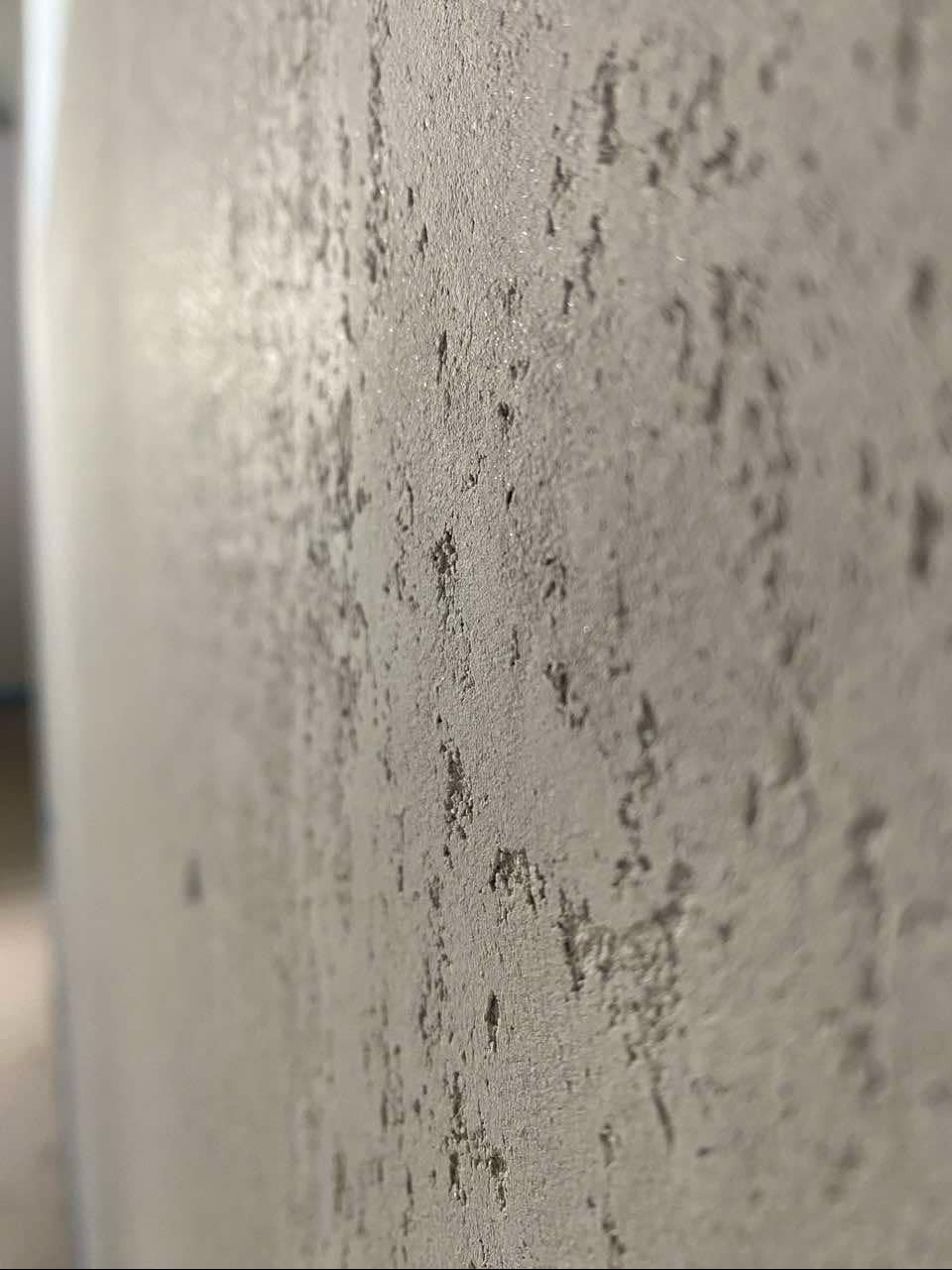 Szara ściana z dekoracyjną, chropowatą strukturą, widoczna z bliskiej perspektywy, z delikatnym połyskiem w górnej części.