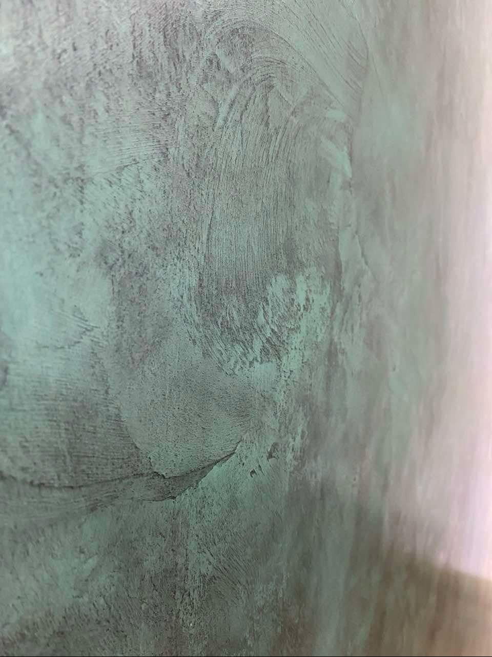 Ściana z dekoracyjnym tynkiem w odcieniach szarości i zieleni, imitującym efekt patyny, z widoczną fakturą i nierównomiernym nałożeniem.