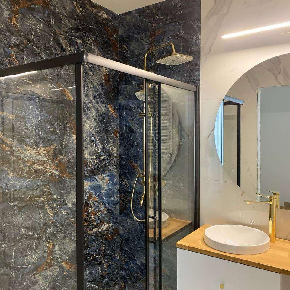 Nowoczesna łazienka z kabiną prysznicową typu walk-in, wyłożona płytkami imitującymi marmur w odcieniach granatu i złota, z armaturą w kolorze szczotkowanego złota i okrągłym lustrem...