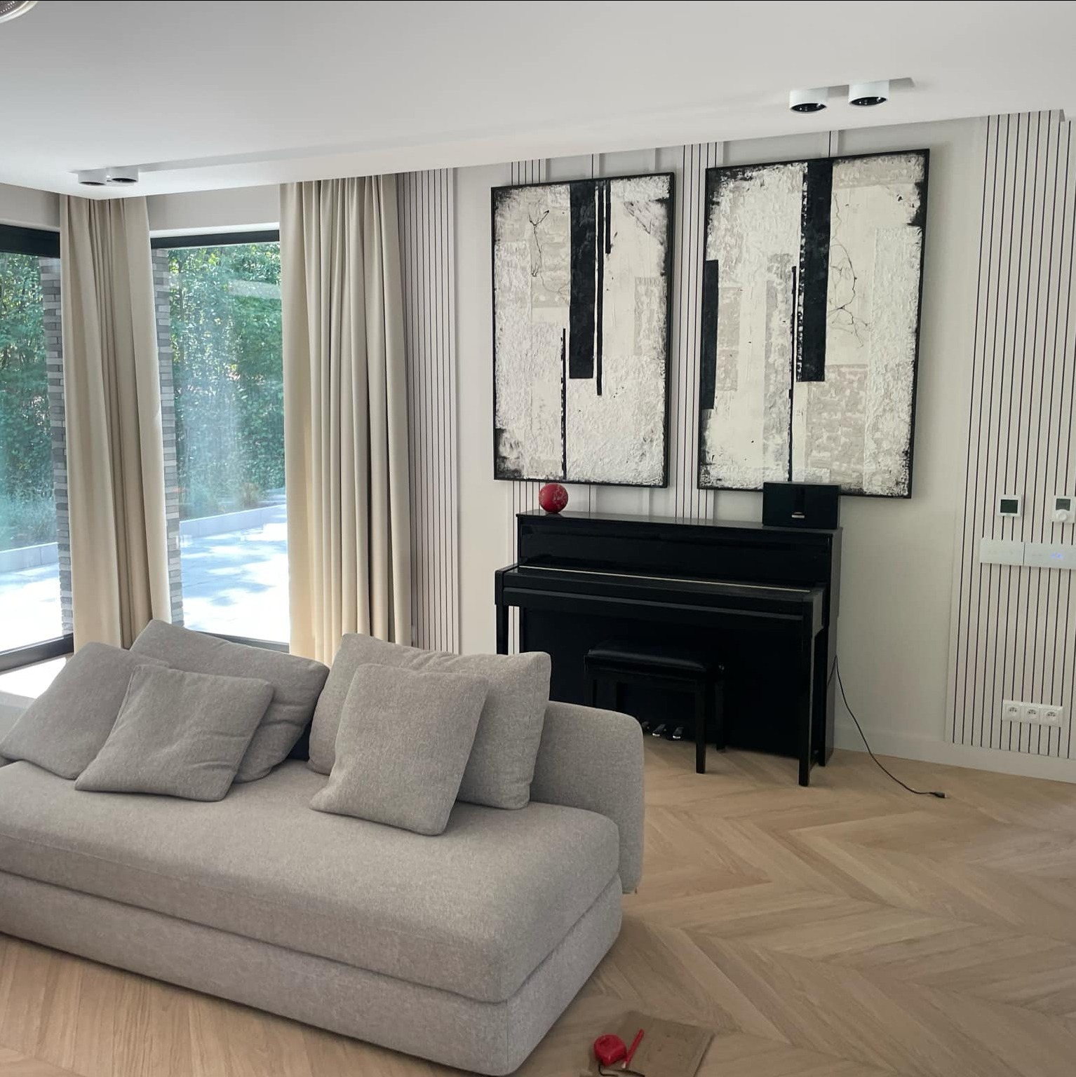 Jasnoszara sofa z poduszkami w salonie z parkietem w jodełkę, czarnym pianinem pod abstrakcyjnymi obrazami i dużym oknem z widokiem na zieleń.