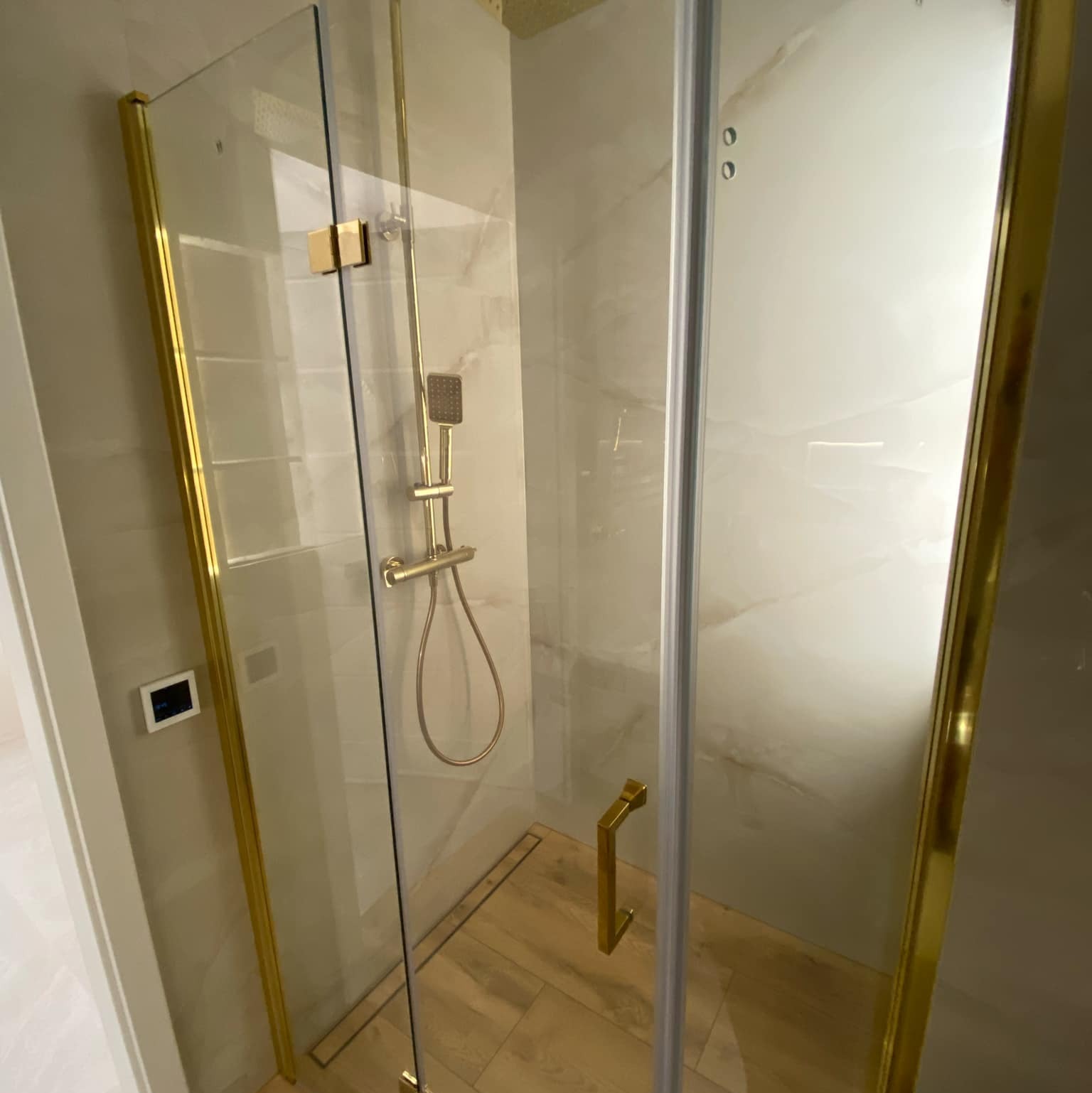 Szklana kabina prysznicowa z złotymi profilami, deszczownicą i odpływem liniowym w podłodze wyłożonej drewnopodobnymi płytkami.