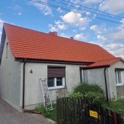 ALLPROFI PLUS - Dom jednorodzinny z nową czerwoną dachówką, widoczna drabina oparta o ścianę, szara elewacja, brązowe rynny i częściowo zasłonięte okno z roletą.