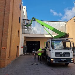 ALLPROFI PLUS - Pracownik na podnośniku koszowym czyści przeszkloną elewację budynku przemysłowego; widoczny znak 'Zakaz Palenia'.