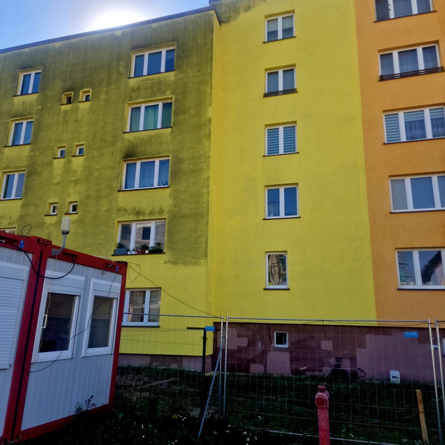 Częściowo oczyszczona elewacja bloku mieszkalnego, widoczny kontrast między brudną i czystą powierzchnią, z kontenerem budowlanym i ogrodzeniem w tle.