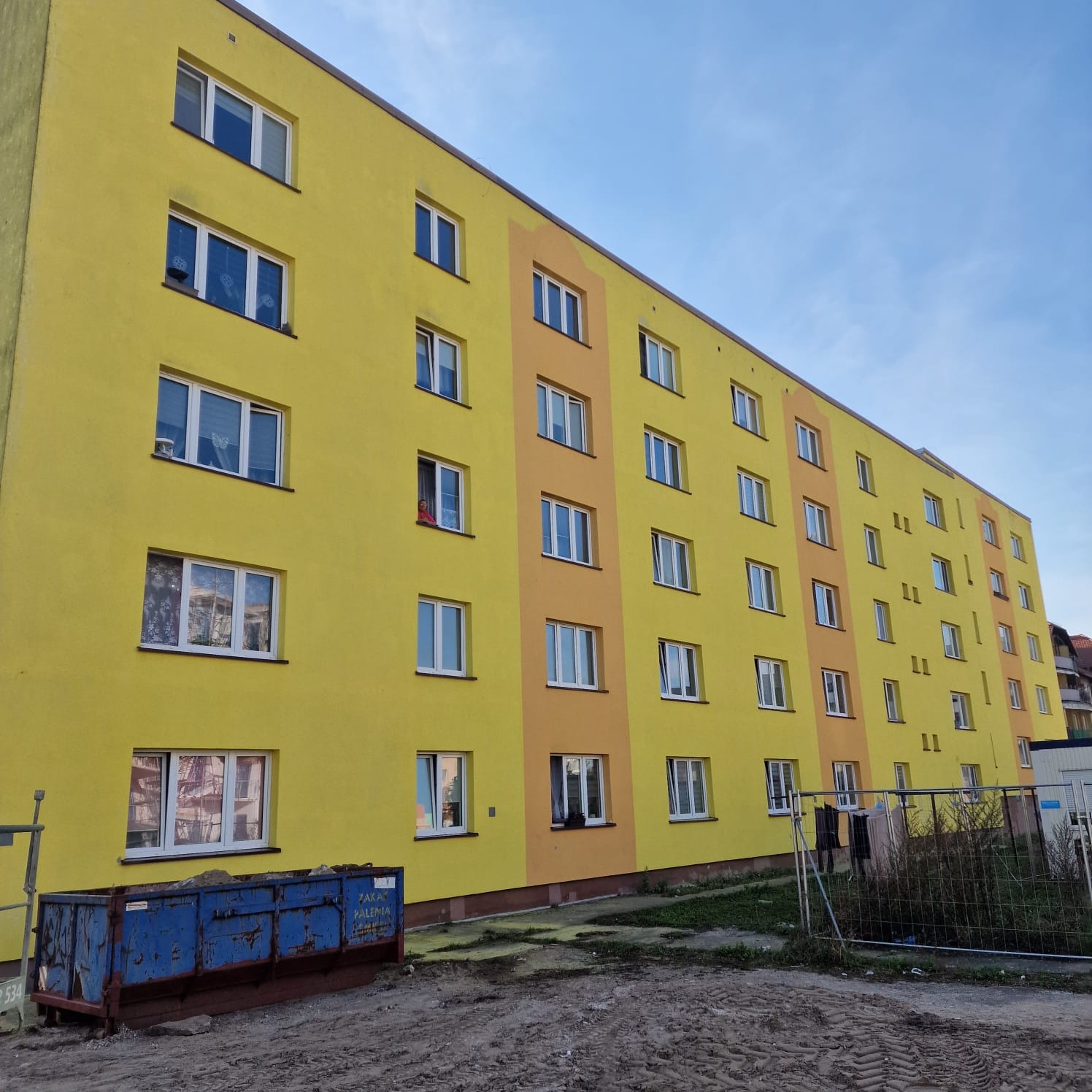 Żółto-pomarańczowy blok mieszkalny po renowacji elewacji, widoczny kontener na gruz i teren budowy.