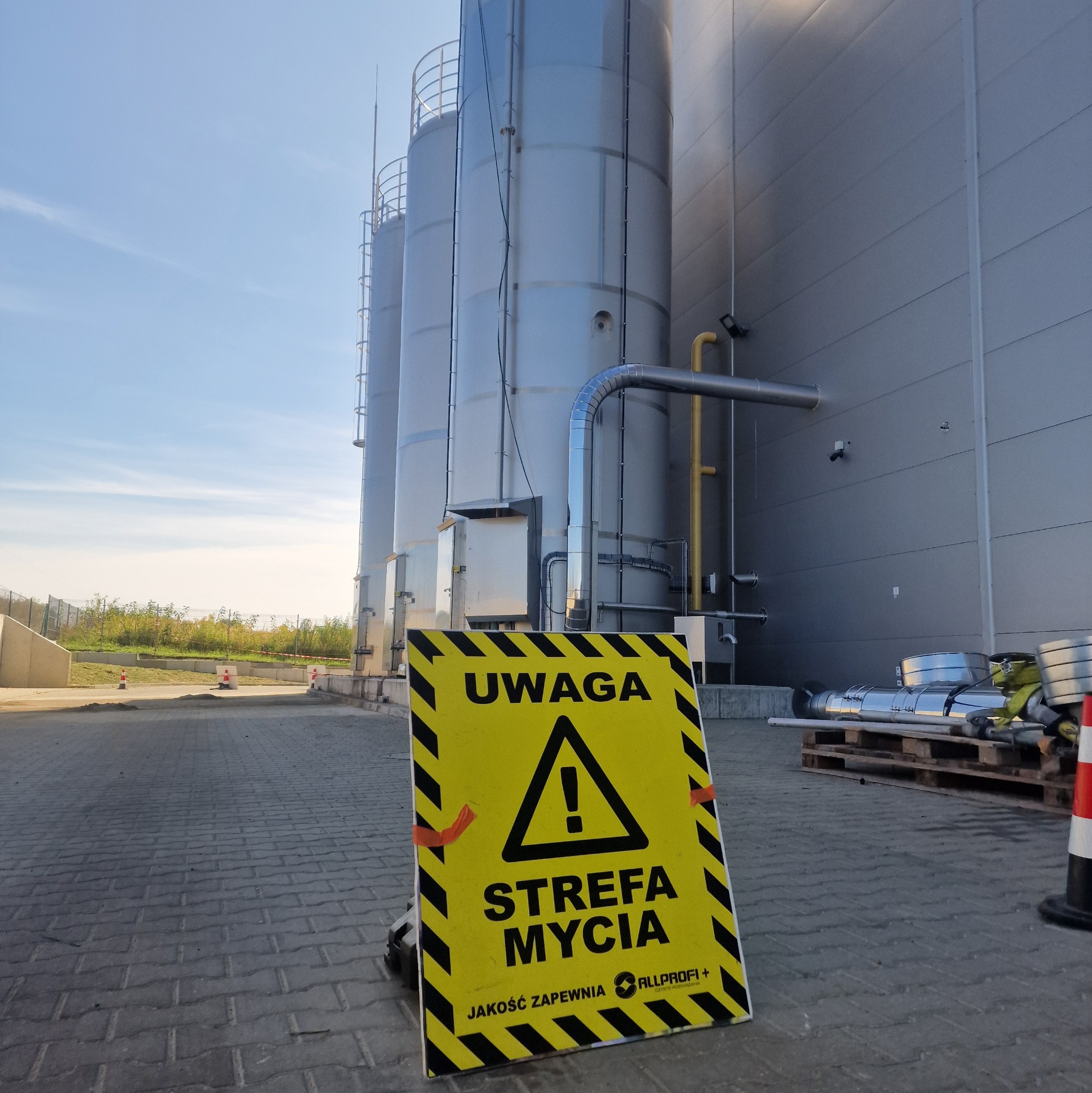 Żółto-czarny znak ostrzegawczy 'Uwaga! Strefa mycia' na kostce brukowej, w tle srebrne silosy przemysłowe i fragment elewacji budynku.