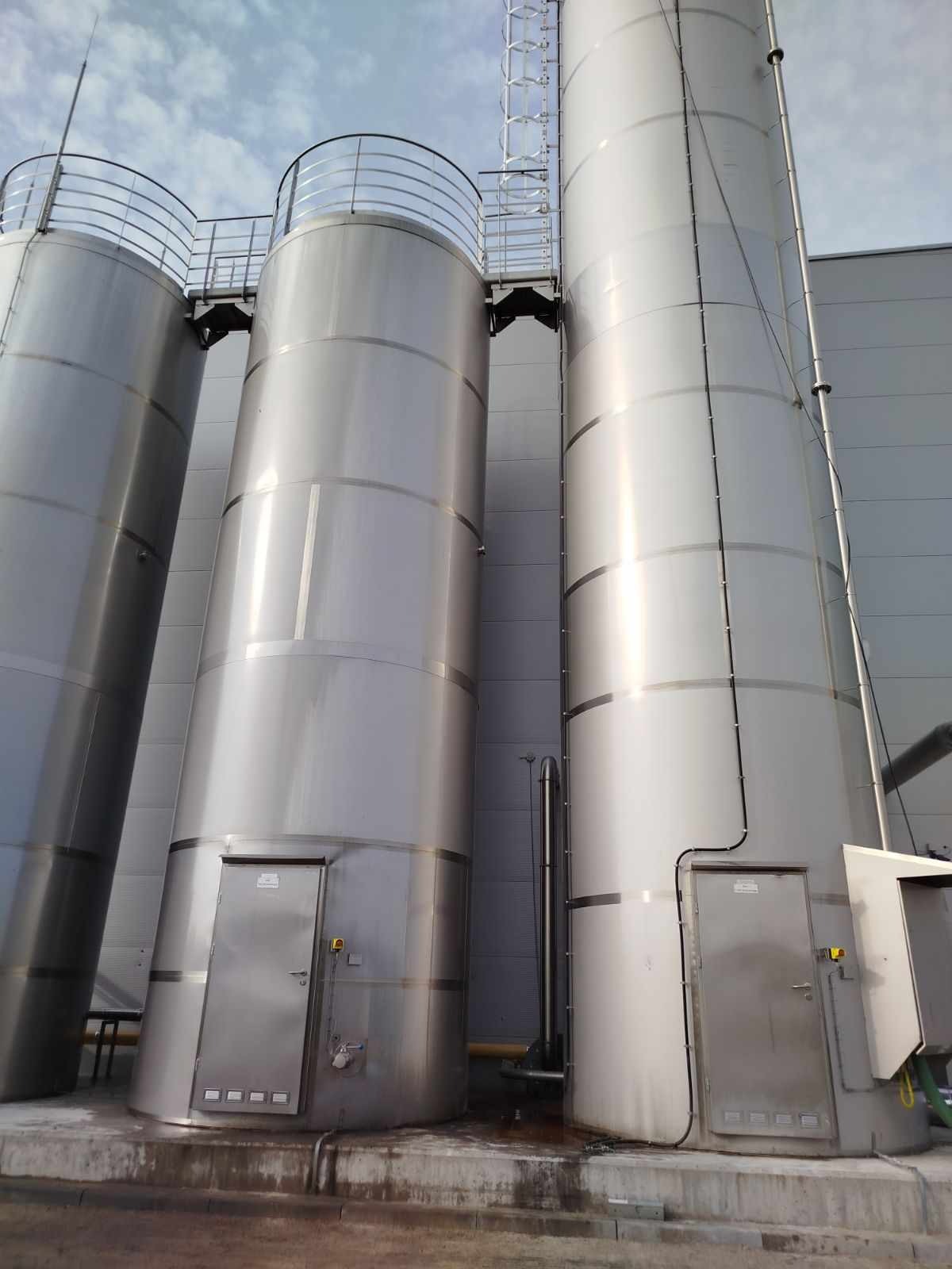 Trzy wysokie, metalowe zbiorniki przemysłowe z drabinkami i platformami, ustawione przy szarym budynku, widoczne ślady zacieków na podstawie.