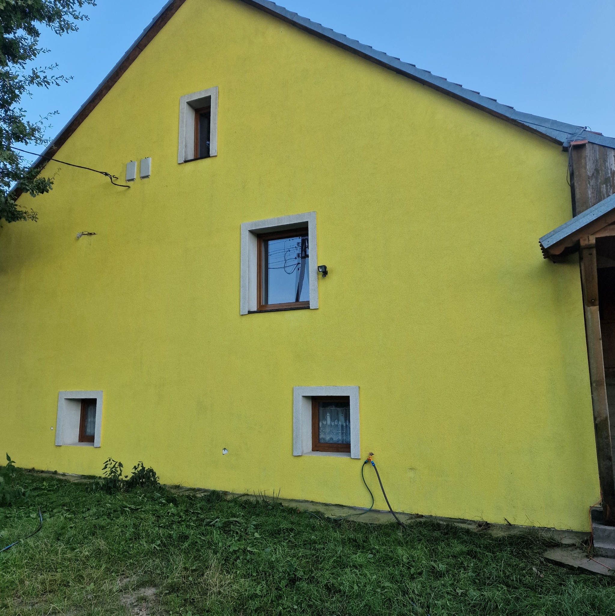 Żółta elewacja budynku z trzema oknami o białych ramach, z kranem i podłączonym wężem ogrodowym przy jednym z okien na parterze. Widoczny fragment dachu i zieleń trawy u podnóża ściany.