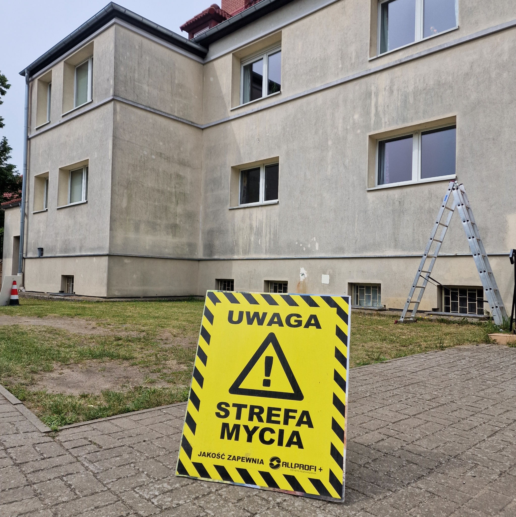 Żółta tablica ostrzegawcza z napisem 'Uwaga Strefa Mycia' przed budynkiem z szarą elewacją i rozstawioną drabiną aluminiową.