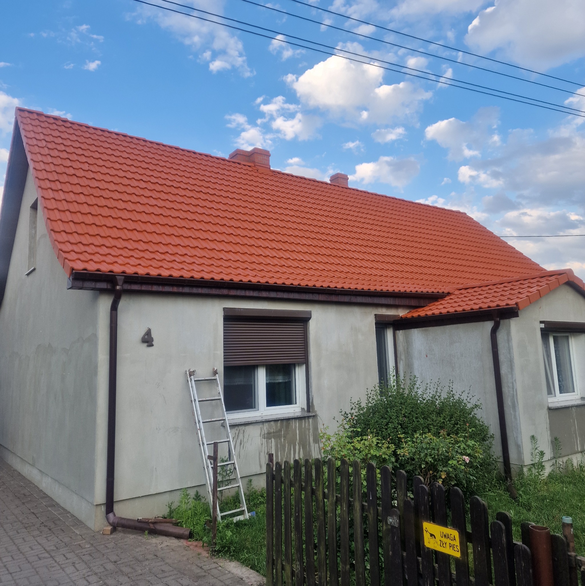 Dom jednorodzinny z nową czerwoną dachówką, widoczna drabina oparta o ścianę, szara elewacja, brązowe rynny i częściowo zasłonięte okno z roletą.
