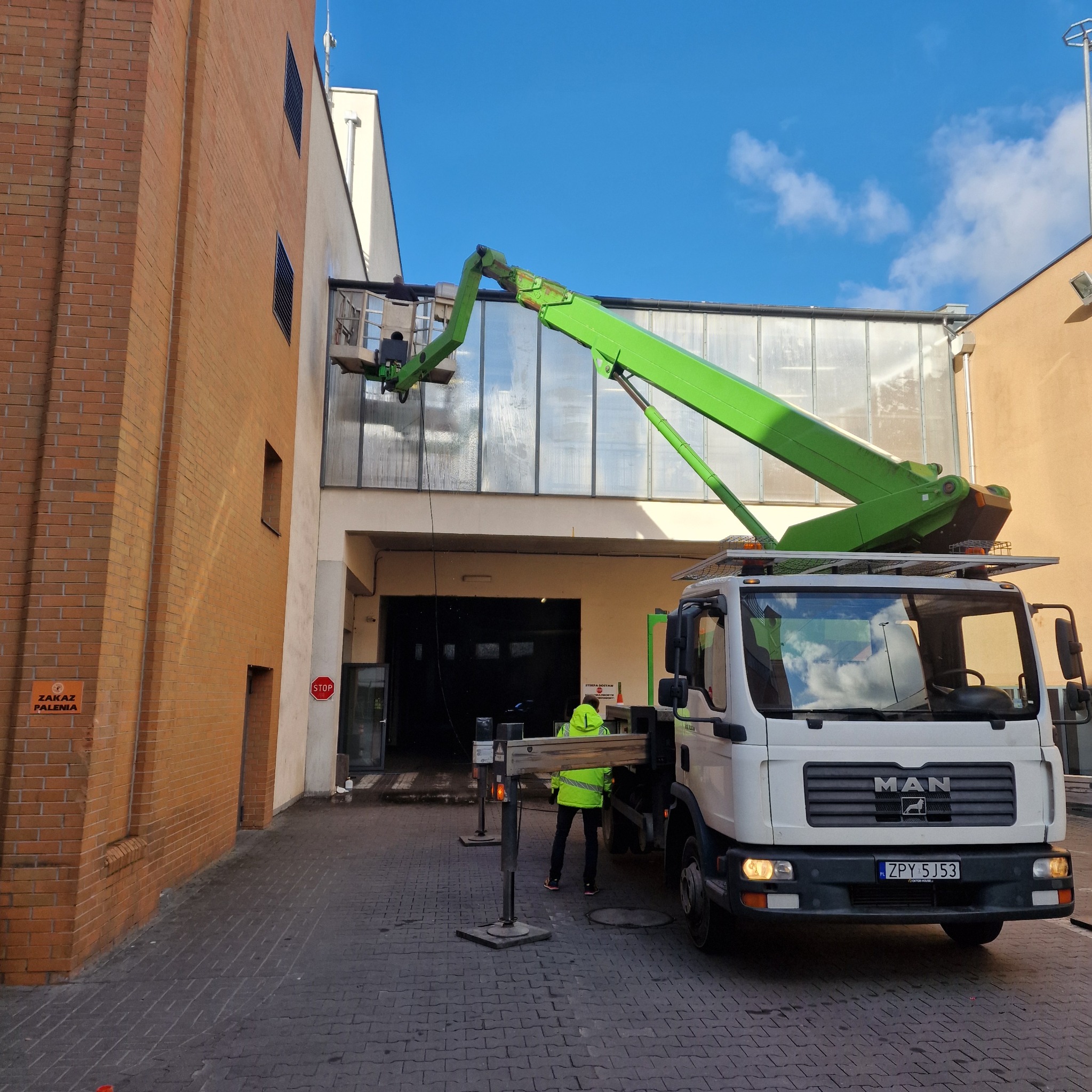 Pracownik na podnośniku koszowym czyści przeszkloną elewację budynku przemysłowego; widoczny znak 'Zakaz Palenia'.