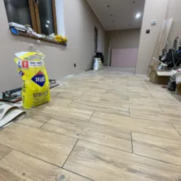 Układanie podłogi z płytek imitujących drewno, widoczny worek z klejem Atlas Geoflex, narzędzia glazurnicze i częściowo ułożona podłoga w długim pomieszczeniu.