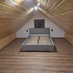 Pokój na poddaszu z drewnianą boazerią na ścianach i suficie, szare łóżko bez materaca na brązowych panelach podłogowych, małe okno w szczycie ściany.