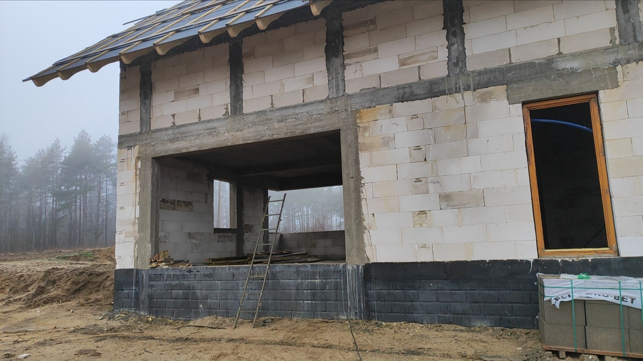 Surowy stan domu jednorodzinnego z betonu komórkowego, widoczne nadproża, częściowo wykonany dach, czarna izolacja fundamentów, okno z drewnianą ramą, w tle zalesiony teren i mgła.