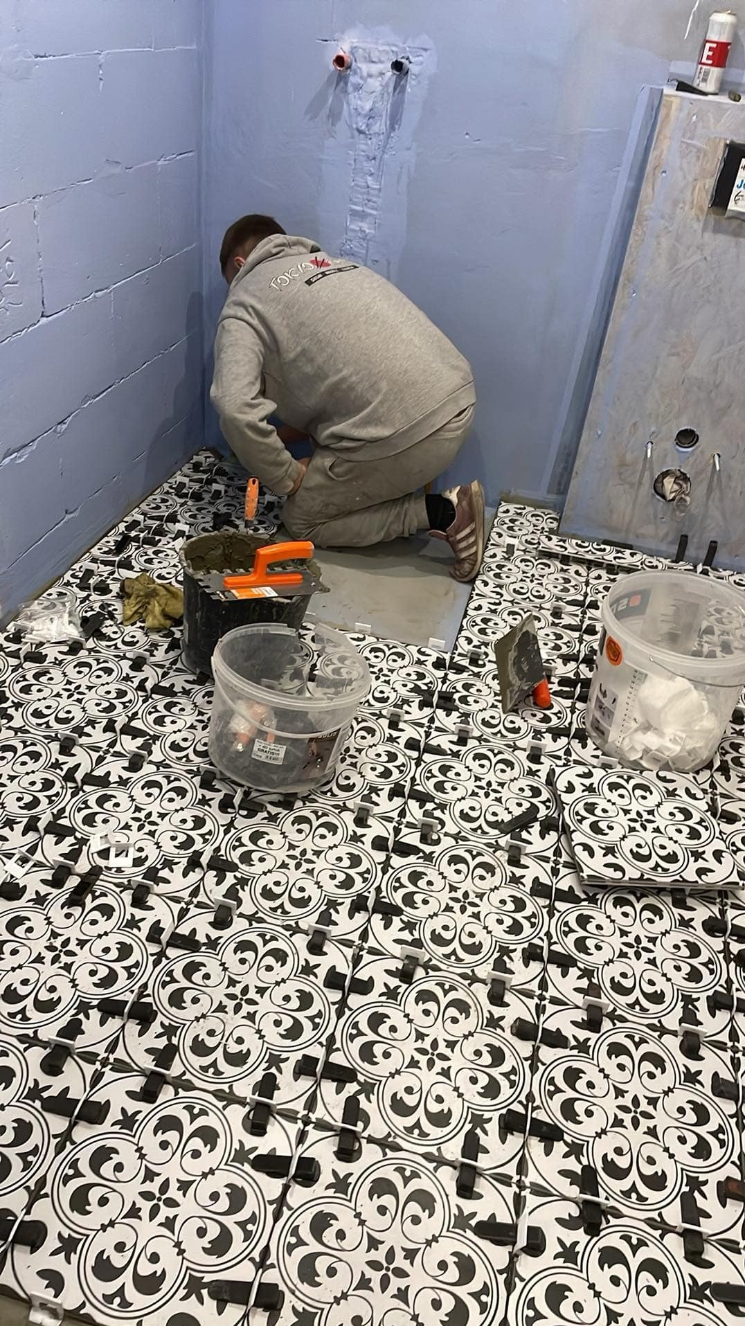 Układanie czarno-białych płytek podłogowych w łazience, widoczne narzędzia, poziomica i plastikowe kliny do fug, ściany przygotowane pod glazurę, pracownik w szarym dresie na kolanach.