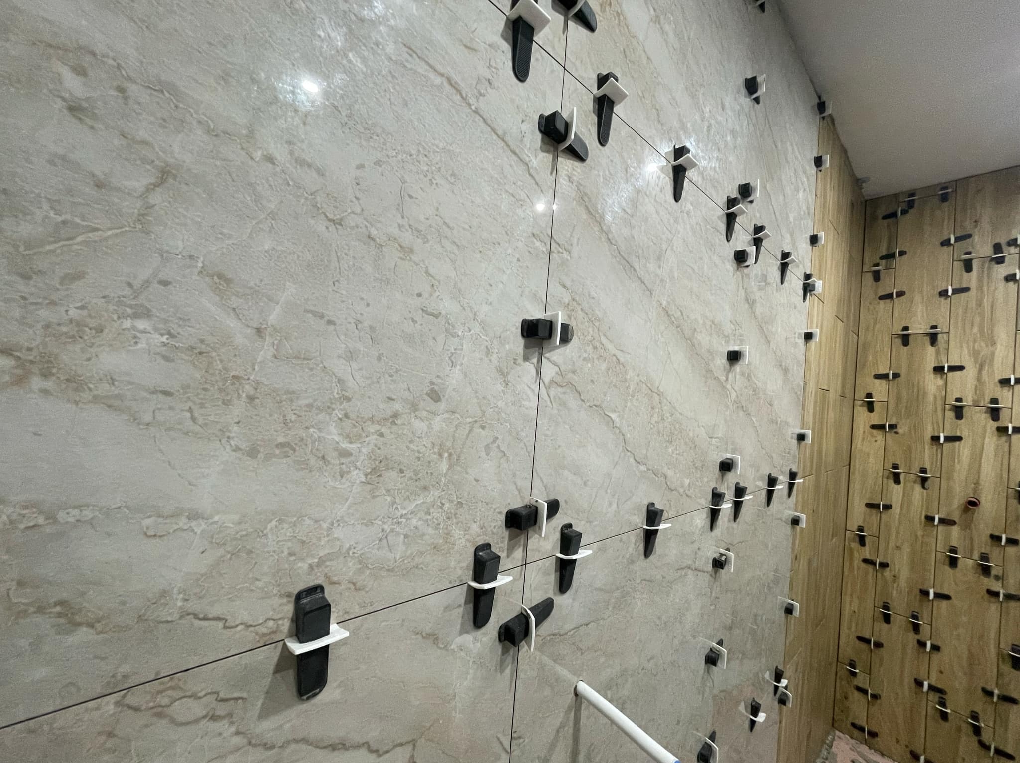 Ściana w trakcie układania płytek imitujących marmur, z użyciem czarnych klipsów poziomujących i białych krzyżyków dystansowych, widoczne fragmenty pionowych sznurków wyznaczających linie.