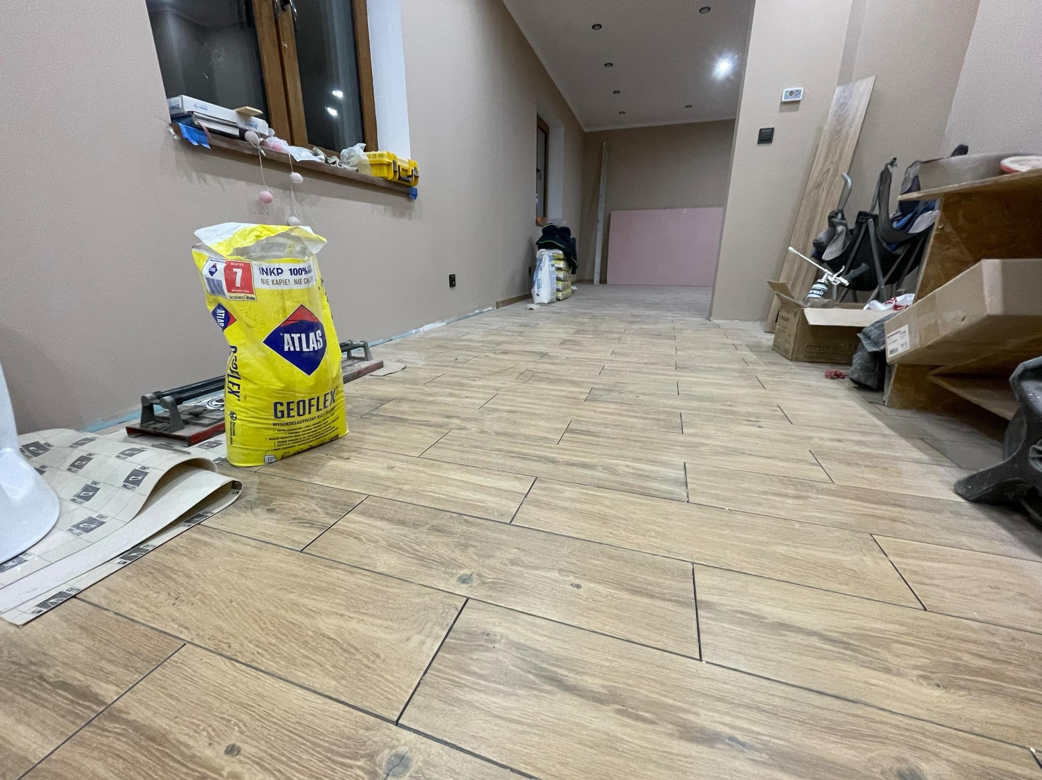Układanie podłogi z płytek imitujących drewno, widoczny worek z klejem Atlas Geoflex, narzędzia glazurnicze i częściowo ułożona podłoga w długim pomieszczeniu.