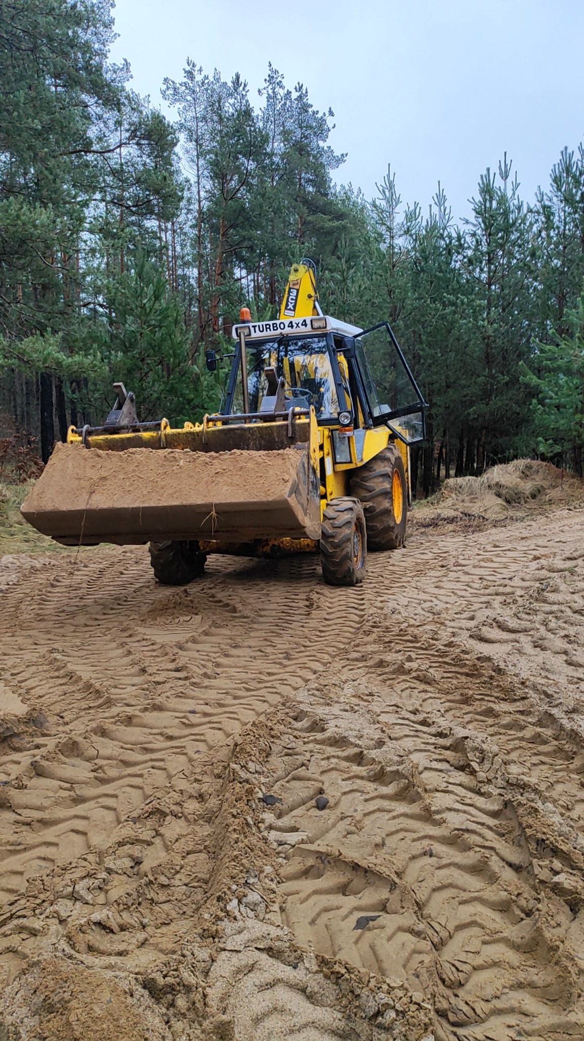 Żółta koparko-ładowarka Turbo 4x4 z otwartymi drzwiami kabiny, stojąca na piaszczystym terenie z widocznymi śladami opon i lasem w tle, pochmurny dzień.