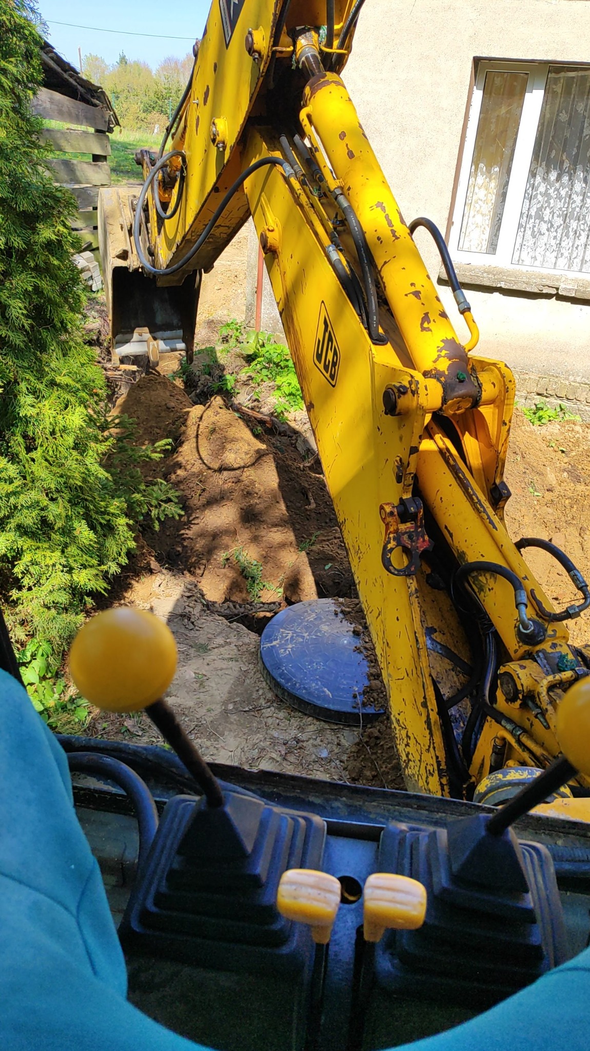 Widok z kabiny żółtej koparki JCB podczas wykopu wąskiego rowu w ziemi, obok zielonego krzewu i studzienki kanalizacyjnej, z widocznymi elementami sterowania koparką.