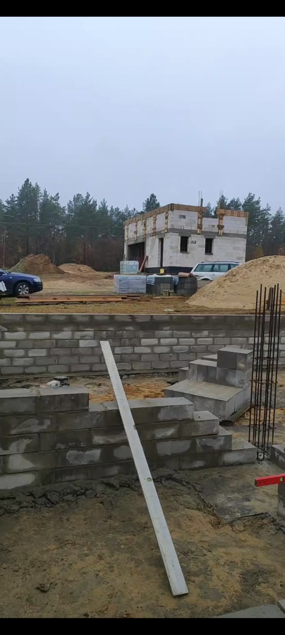 Wznoszenie fundamentów domu z bloczków betonowych, widoczny poziomica, zbrojenie, w tle budynek w stanie surowym otwartym, samochody i drzewa.
