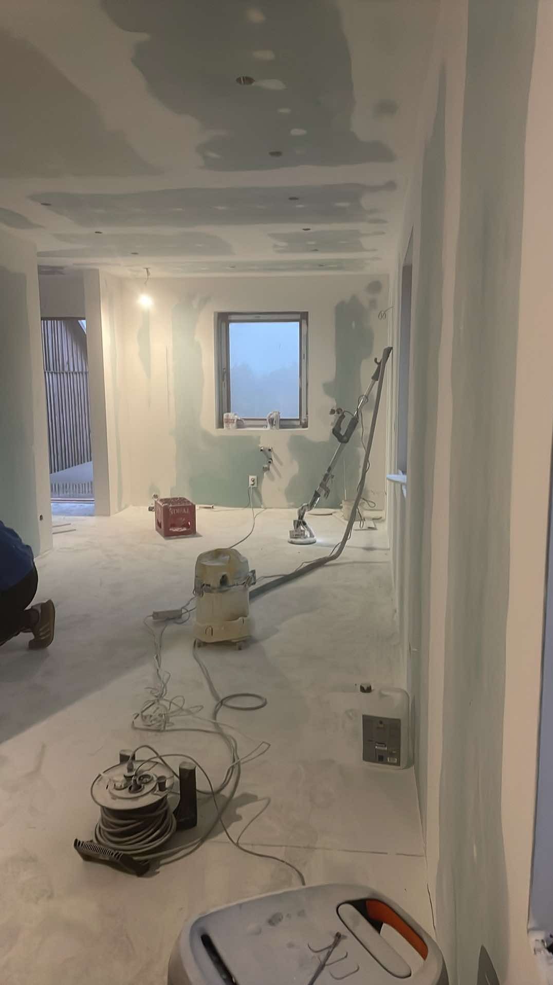 Pokój w trakcie szpachlowania ścian i sufitu, widoczna szlifierka do gipsu z odkurzaczem przemysłowym i zwinięty przedłużacz na podłodze, ściany przygotowane do malowania.