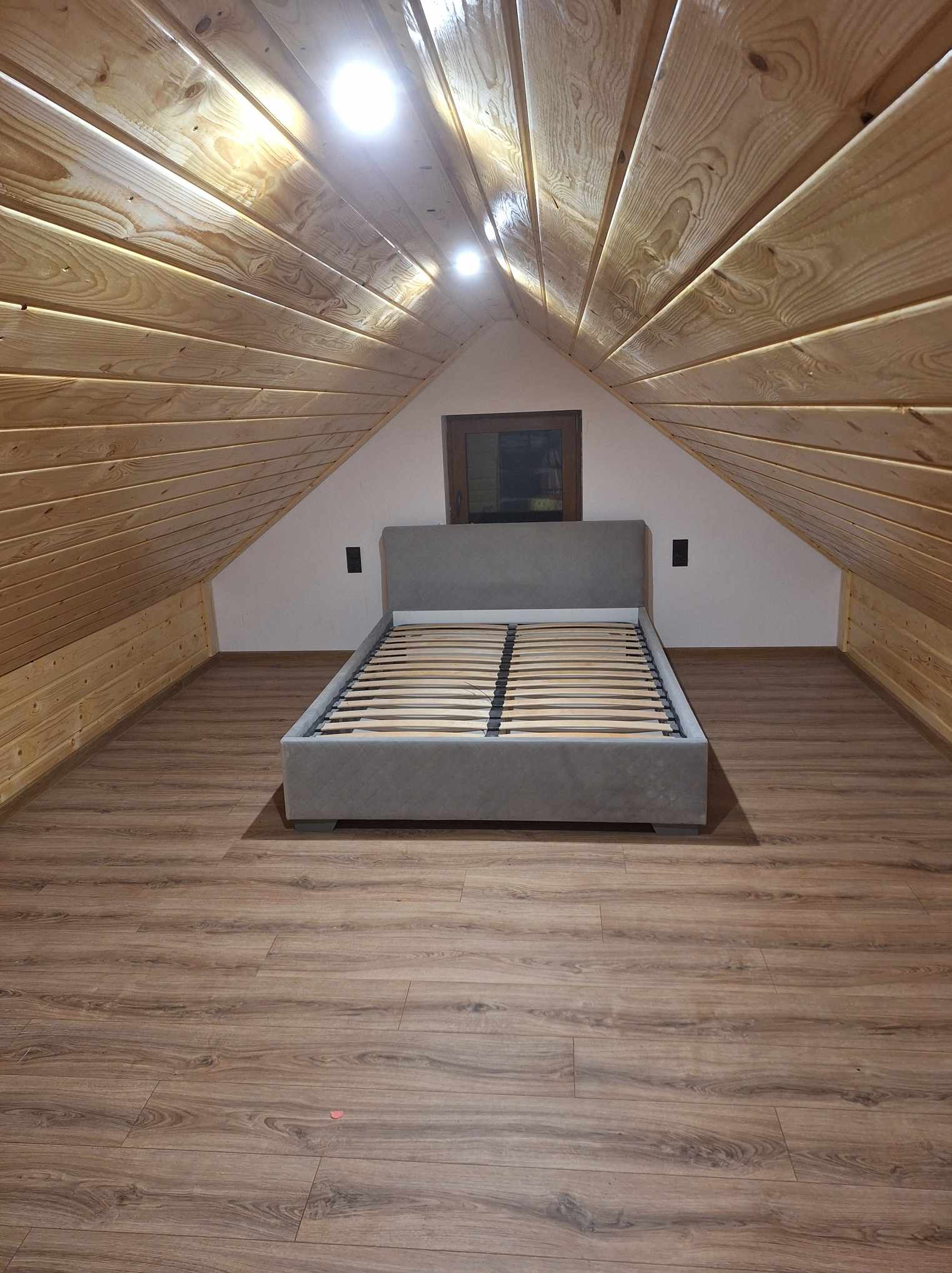 Pokój na poddaszu z drewnianą boazerią na ścianach i suficie, szare łóżko bez materaca na brązowych panelach podłogowych, małe okno w szczycie ściany.