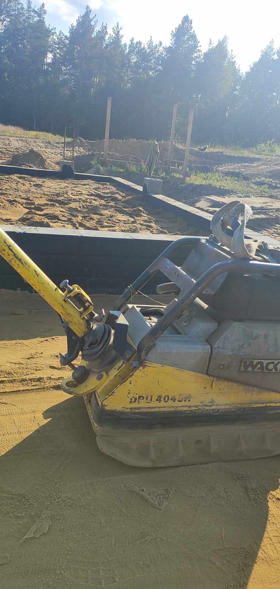 Żółta zagęszczarka Wacker DPU4045H ubija piasek w wykopie pod fundamenty, w tle widoczne drewniane szalunki i las.