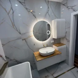 Nowoczesna łazienka z marmurowymi płytkami, okrągłym lustrem z podświetleniem LED, białą umywalką nablatową na drewnianym blacie i białą szafką wiszącą